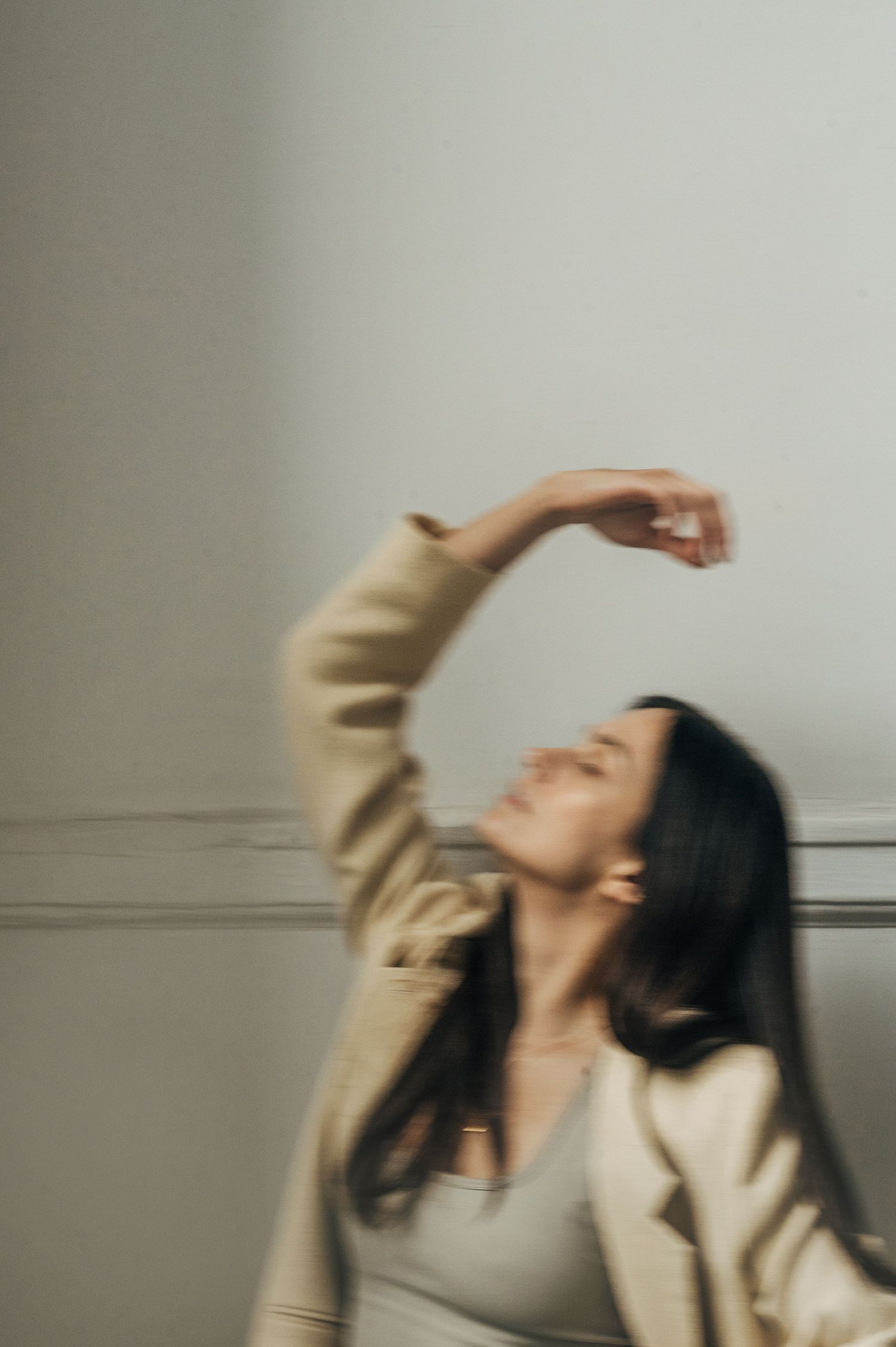 Dancing figure in beige blazer creates artistic motion blur against minimalist white wall.