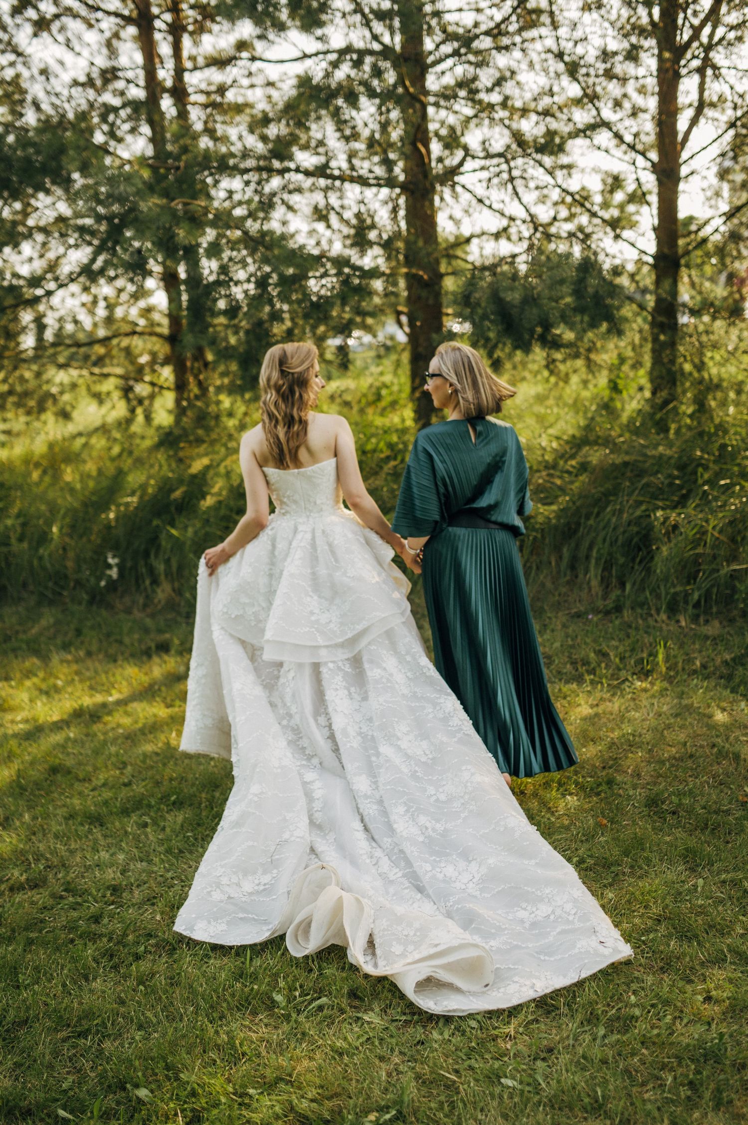 Bride in white gown holds hands with woman in green dress walking through lush forest setting.