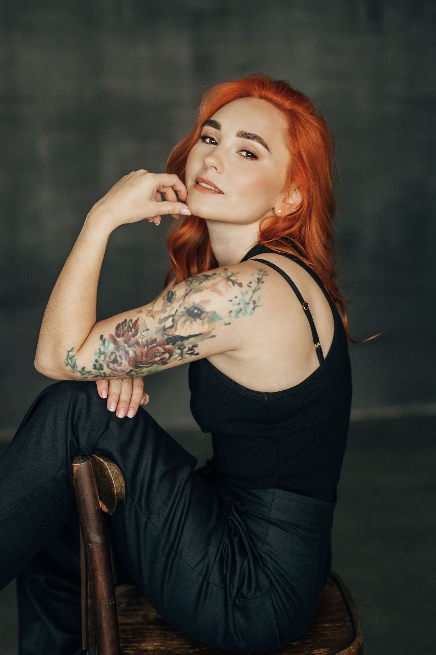 Woman with red hair and tattoos poses in black dress against dark background in artistic portrait series.