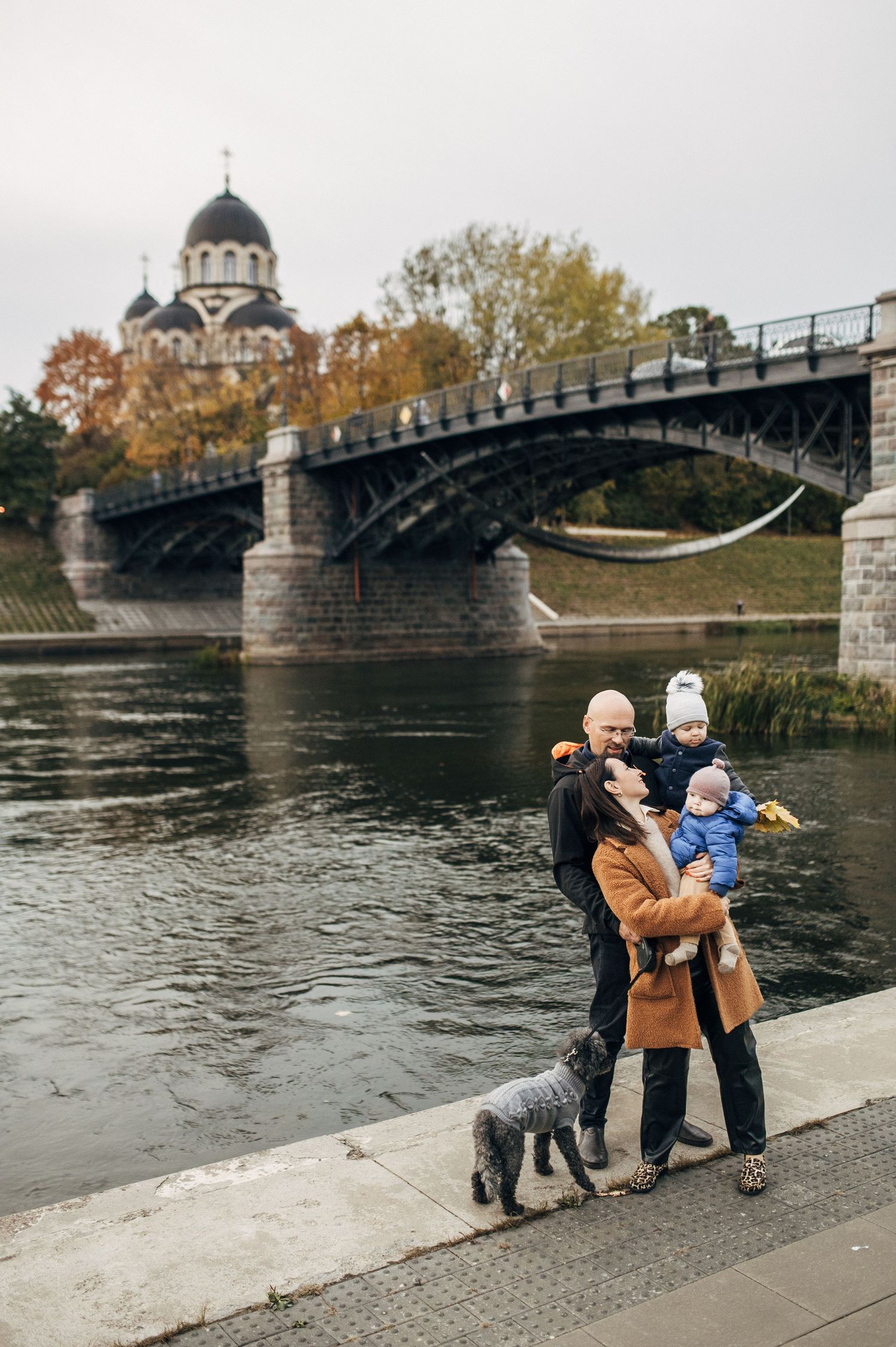 Family enjoys autumn day by riverbank with historic bridge and domed building in background.