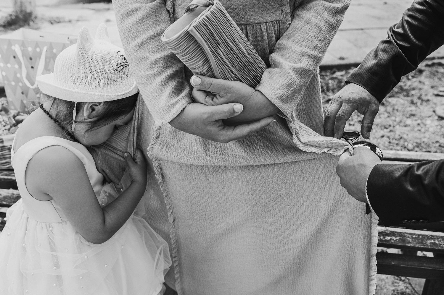 Young girl in white dress hiding face beside adult holding book at outdoor event.