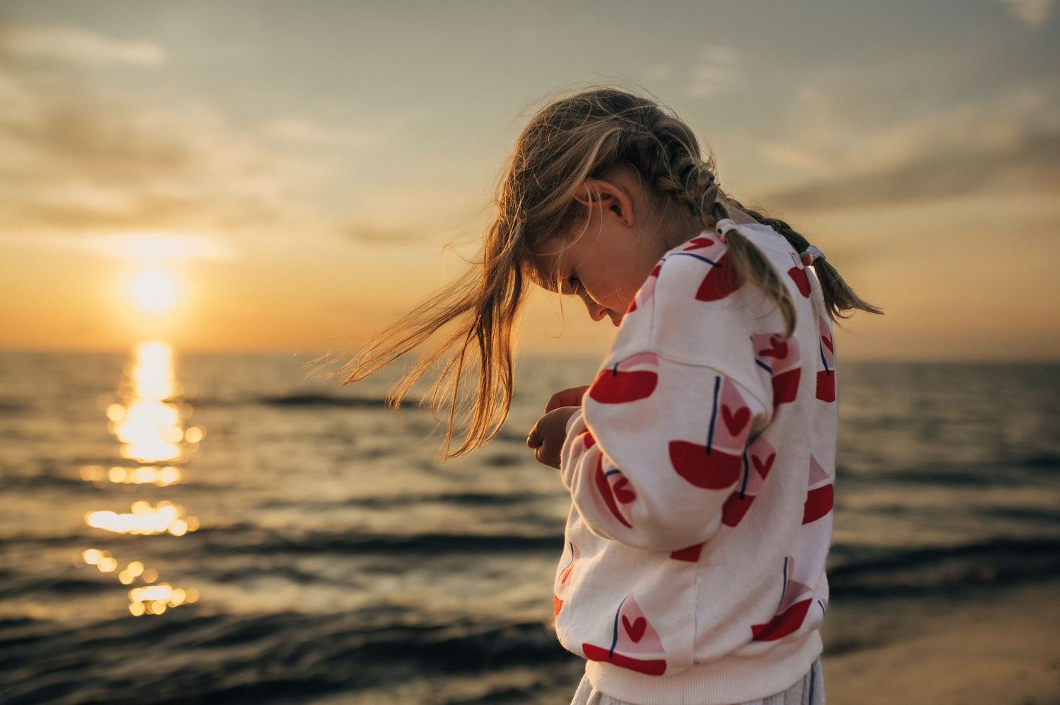 Young girl in red heart hoodie stands on beach at golden sunset, hair blowing in breeze over ocean.