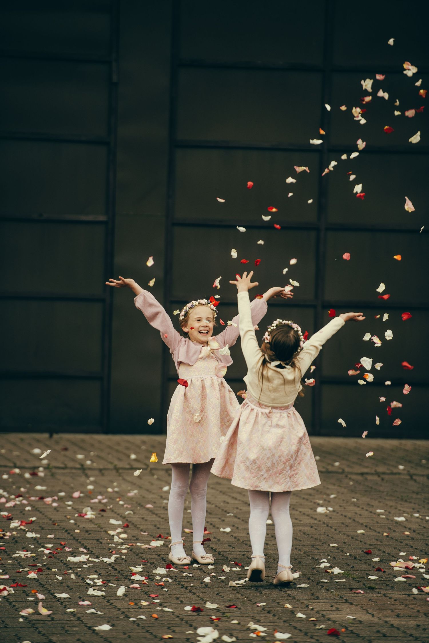 Two joyful girls in pink dresses tossing colorful confetti petals in the air outdoors.