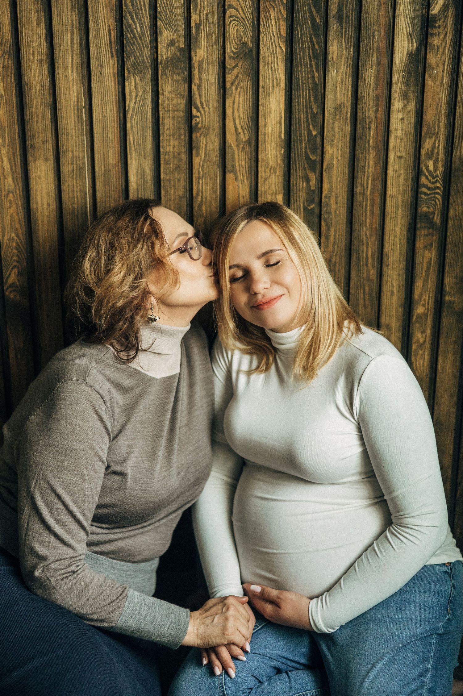 Intimate moment shared between two women during maternity photoshoot against wooden backdrop.