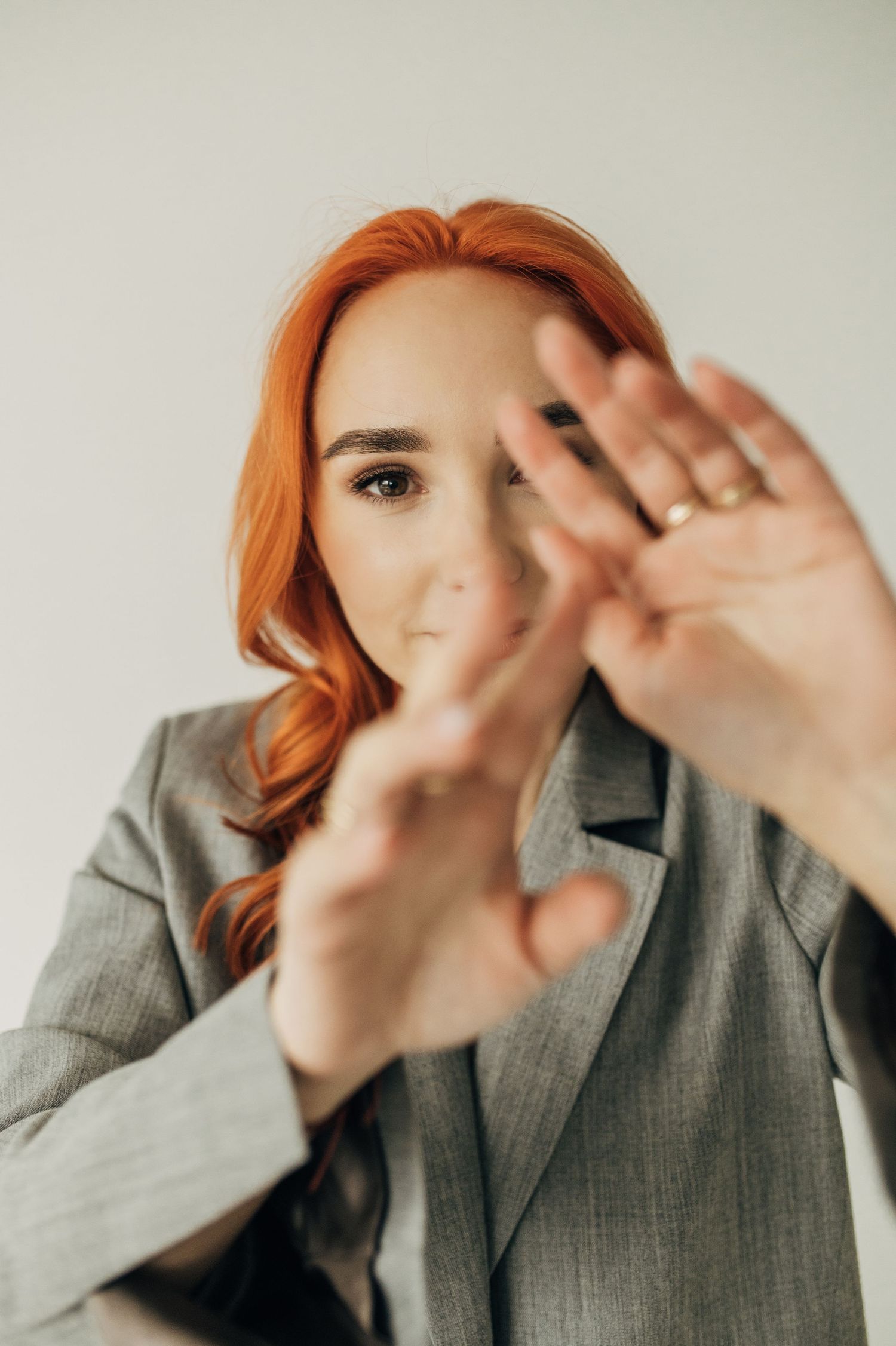 Figure in grey blazer makes expressive hand gestures against light background.