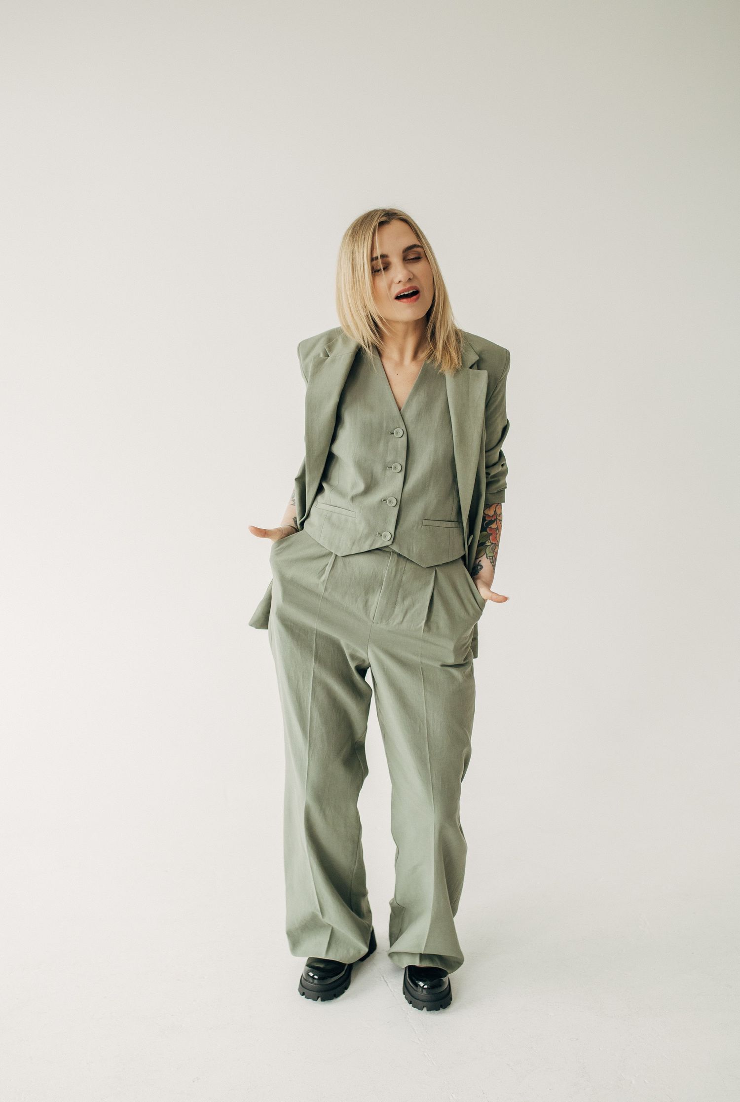 Woman in oversized sage green pantsuit poses against white background.