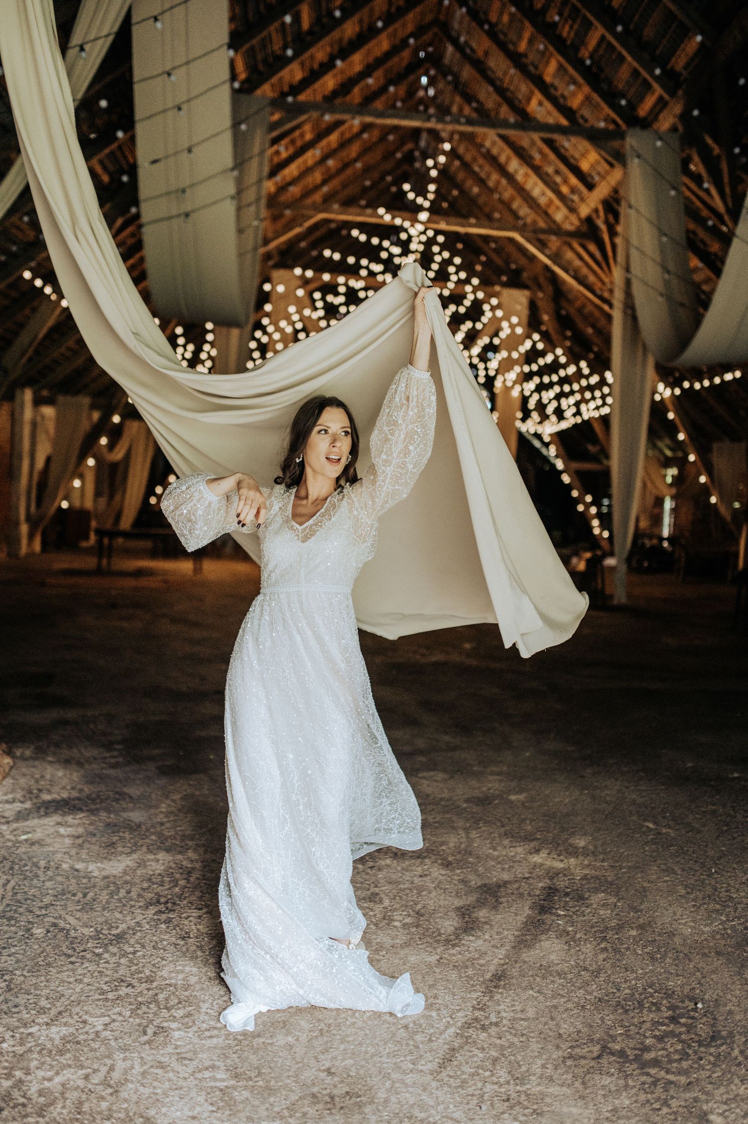 A bride twirls in a white dress under twinkling lights in a rustic barn venue.