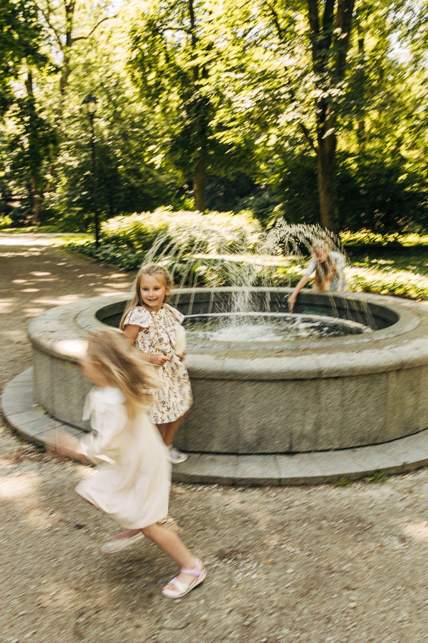 Two young girls in dresses play and run around a stone fountain in a sunny park.