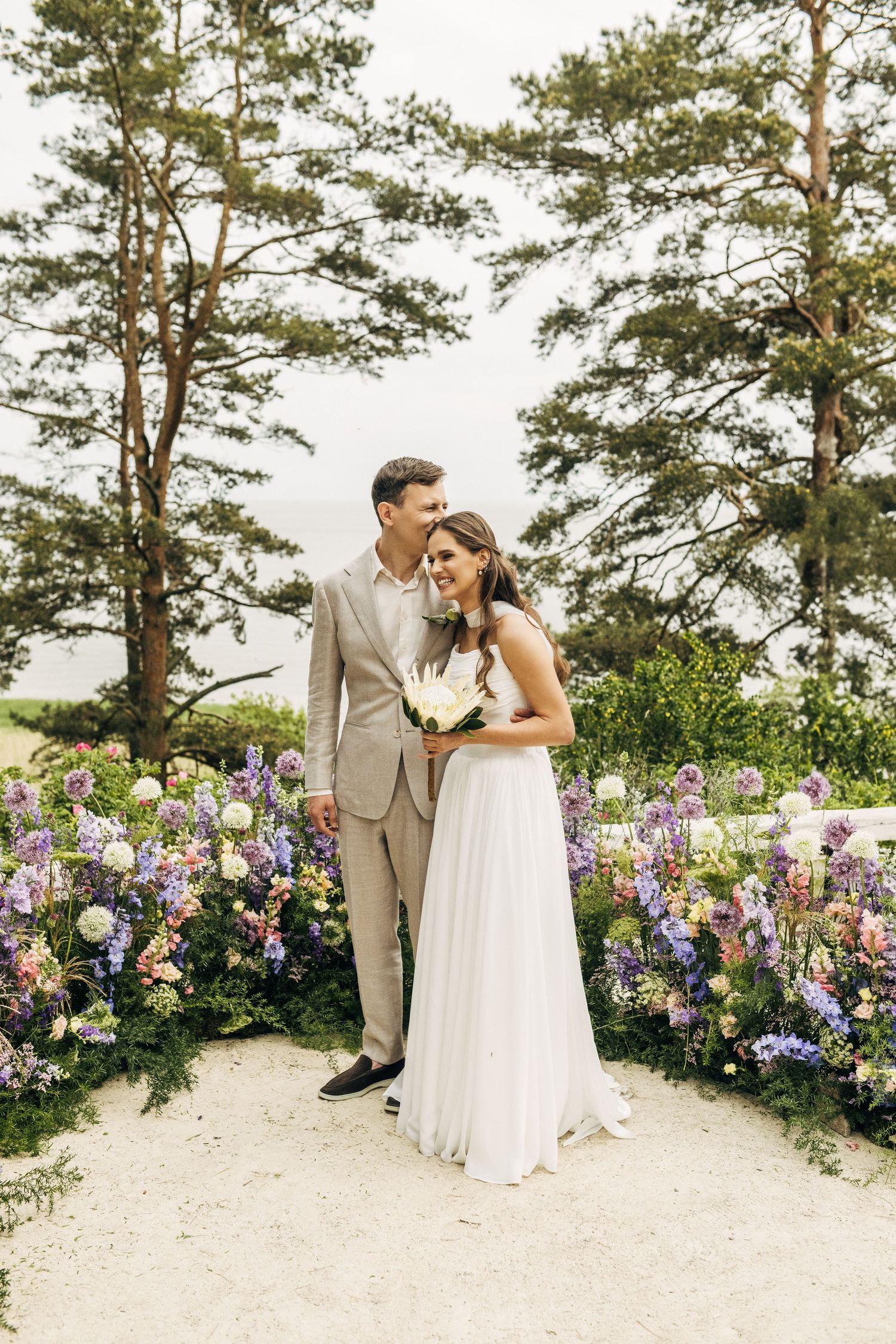A romantic wedding couple shares intimate moments in a garden path lined with purple delphiniums and pine trees.