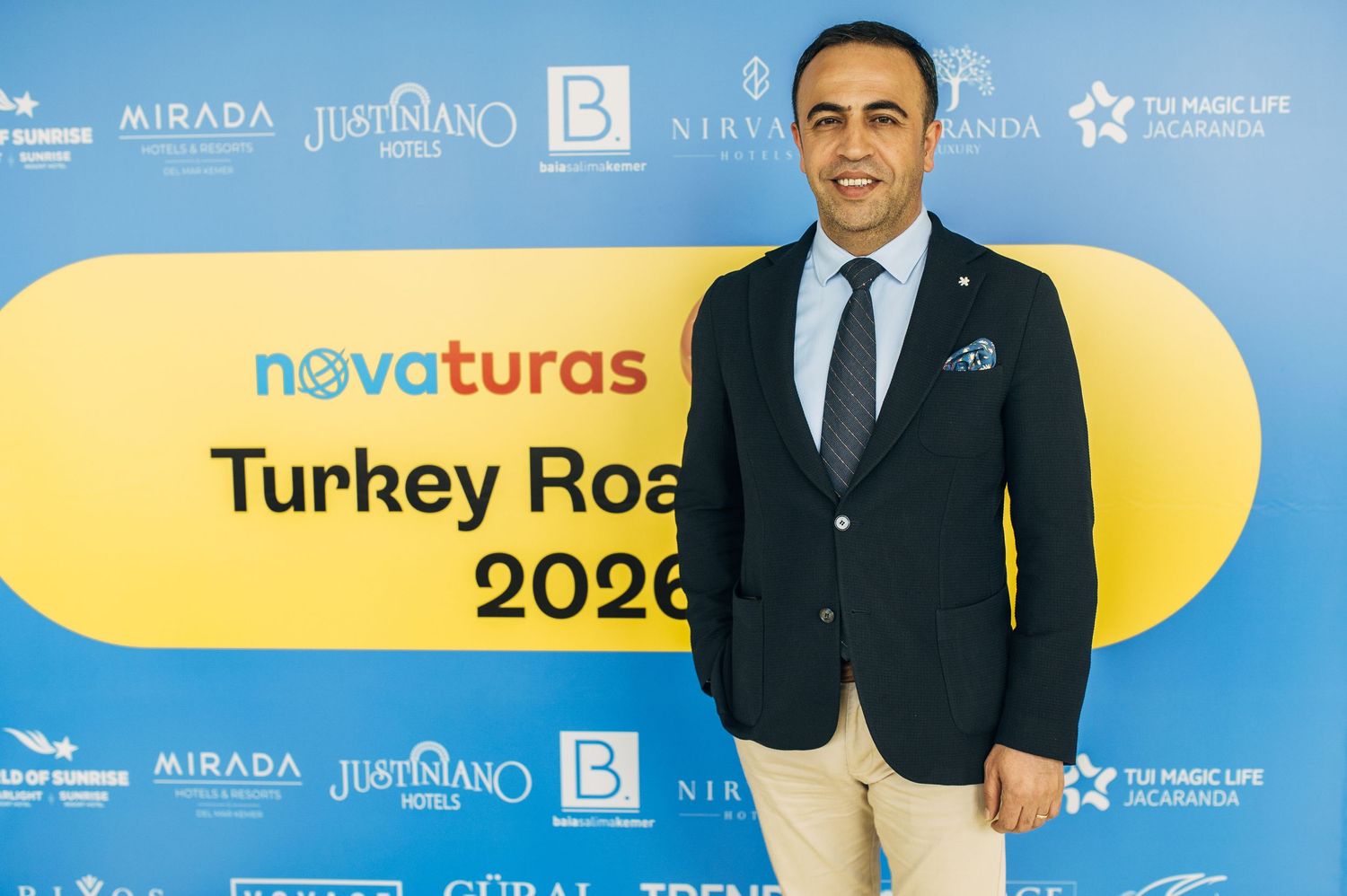 A person in a navy suit jacket and khaki pants stands in front of a yellow backdrop with corporate event branding.