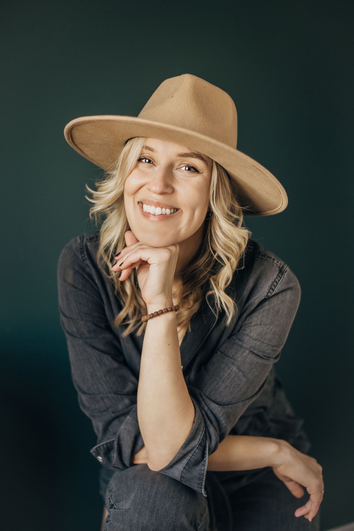 Professional studio portrait with dark backdrop featuring someone in a brown felt hat and charcoal button-down shirt.