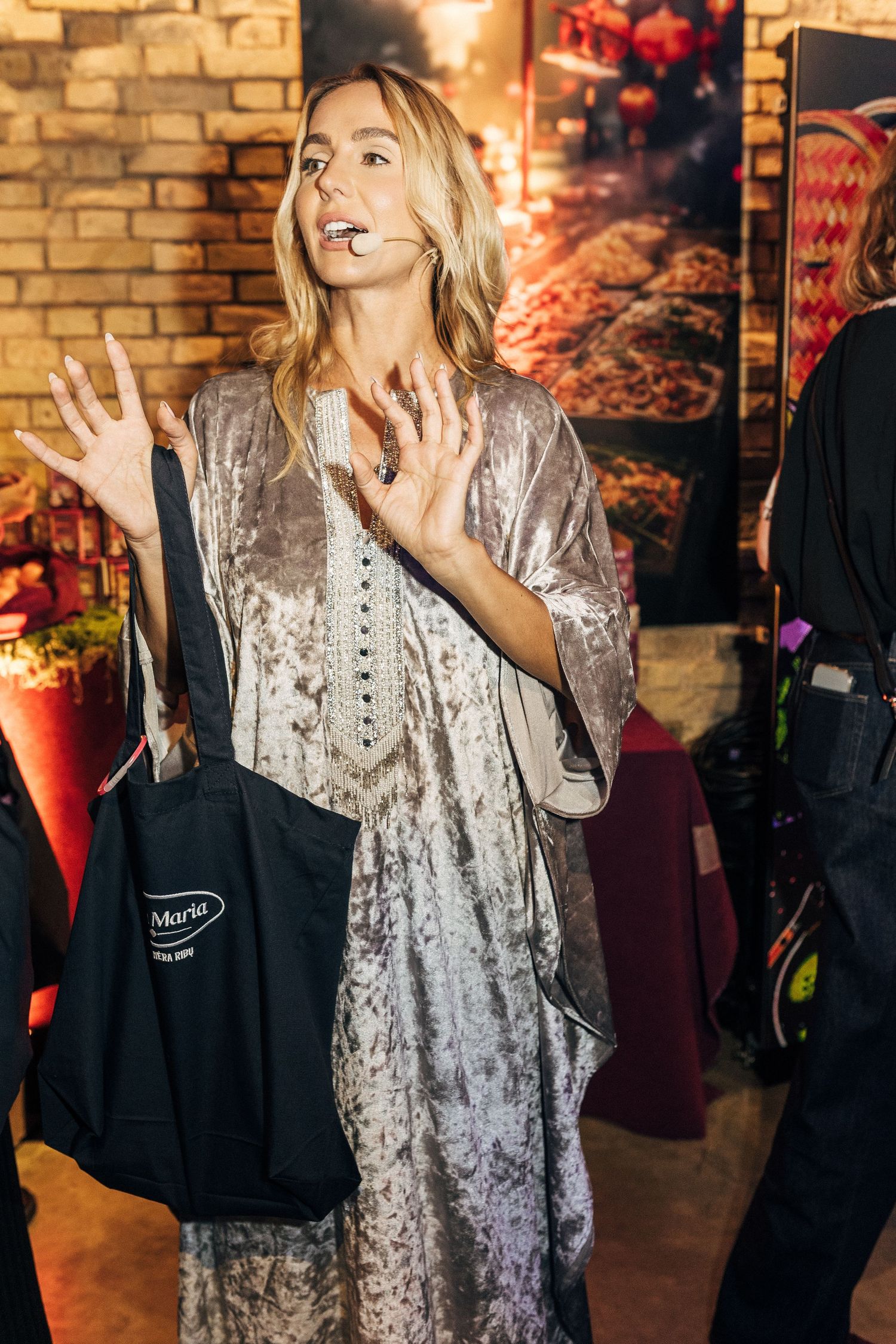 A person in a silver sequined dress gestures animatedly against a brick wall background.
