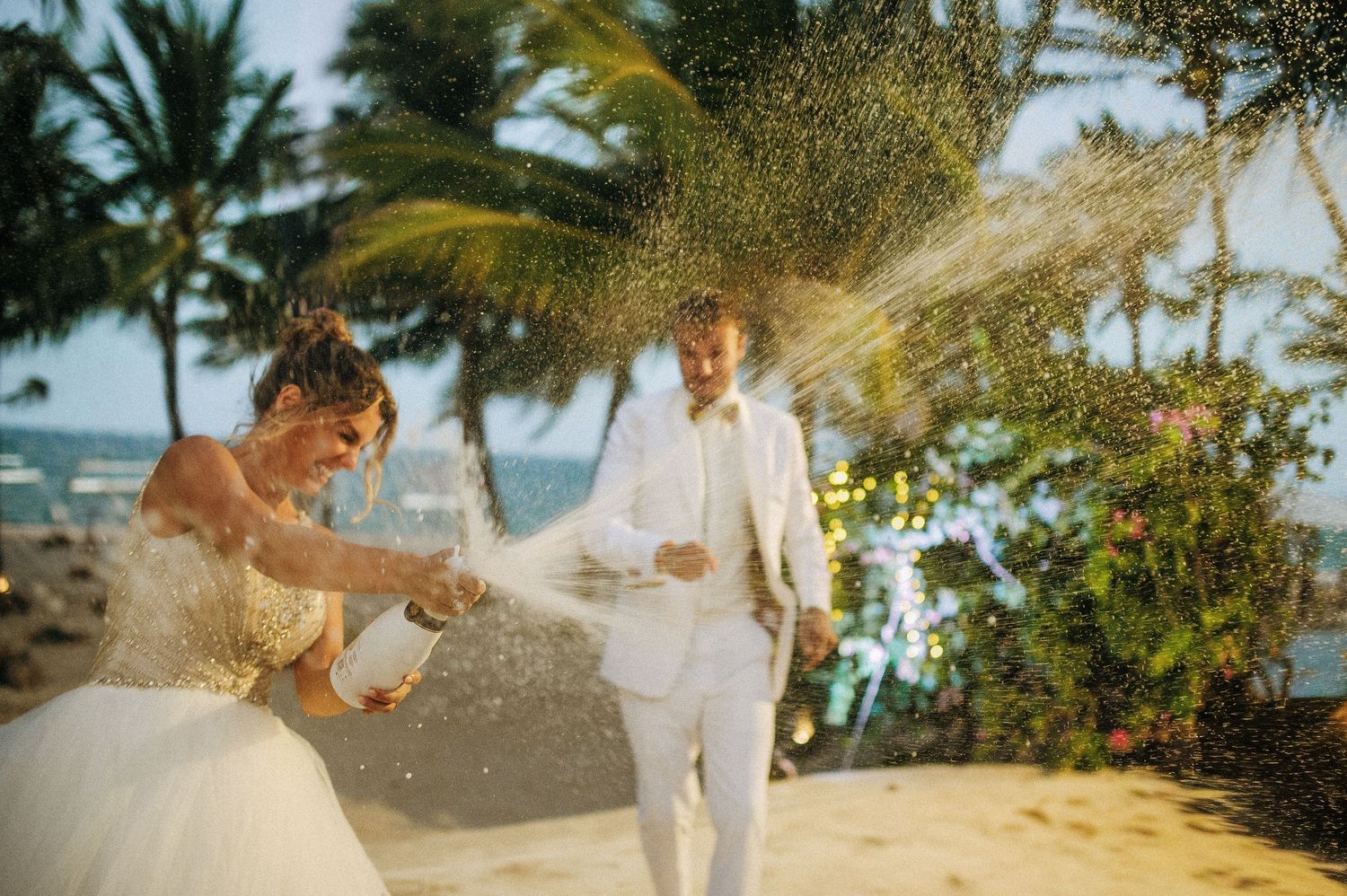 Newlyweds celebrate on a sandy beach as golden sunlight filters through palm trees.