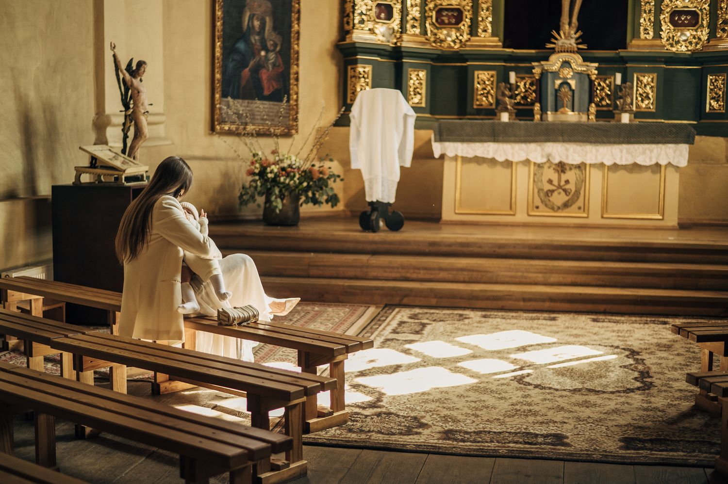 Woman in white dress sits alone on church pew in ornate Catholic chapel with golden altar.