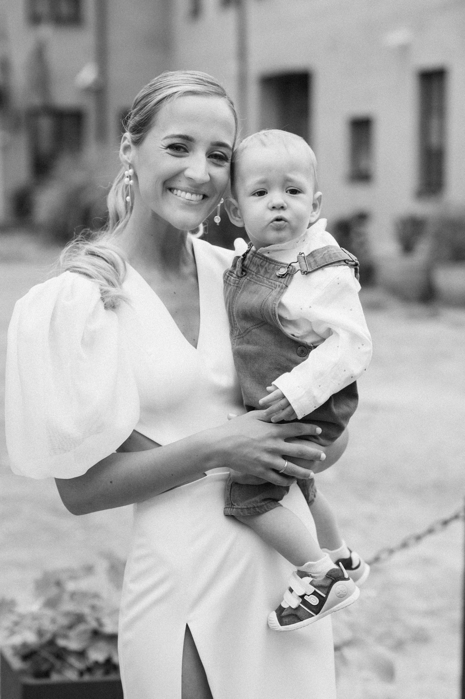 Smiling bride in white dress with puff sleeves holding a baby boy in overalls, black and white portrait.