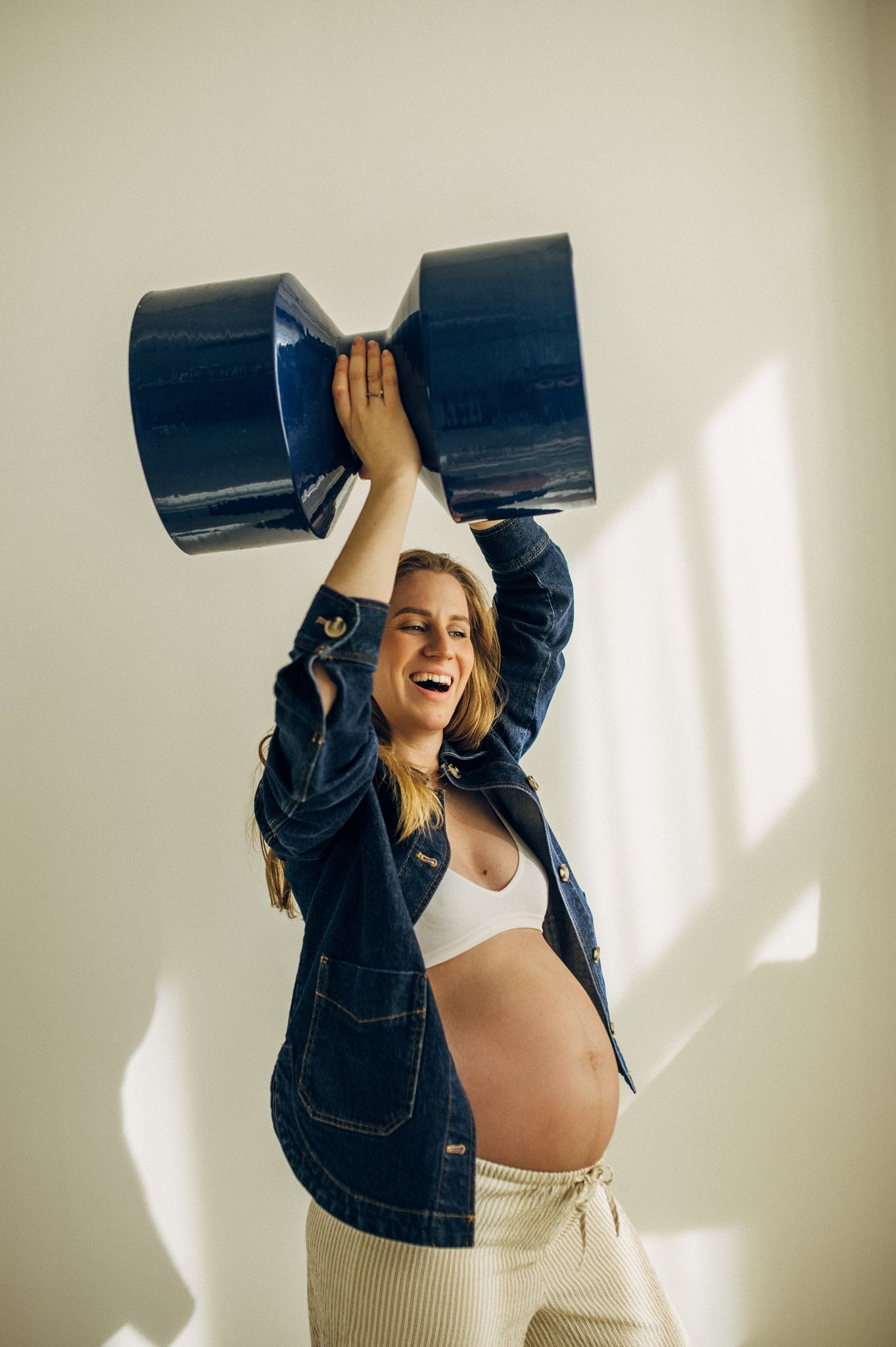 Happy pregnant woman in denim jacket lifting blue pots overhead, laughing while showing her baby bump indoors.
