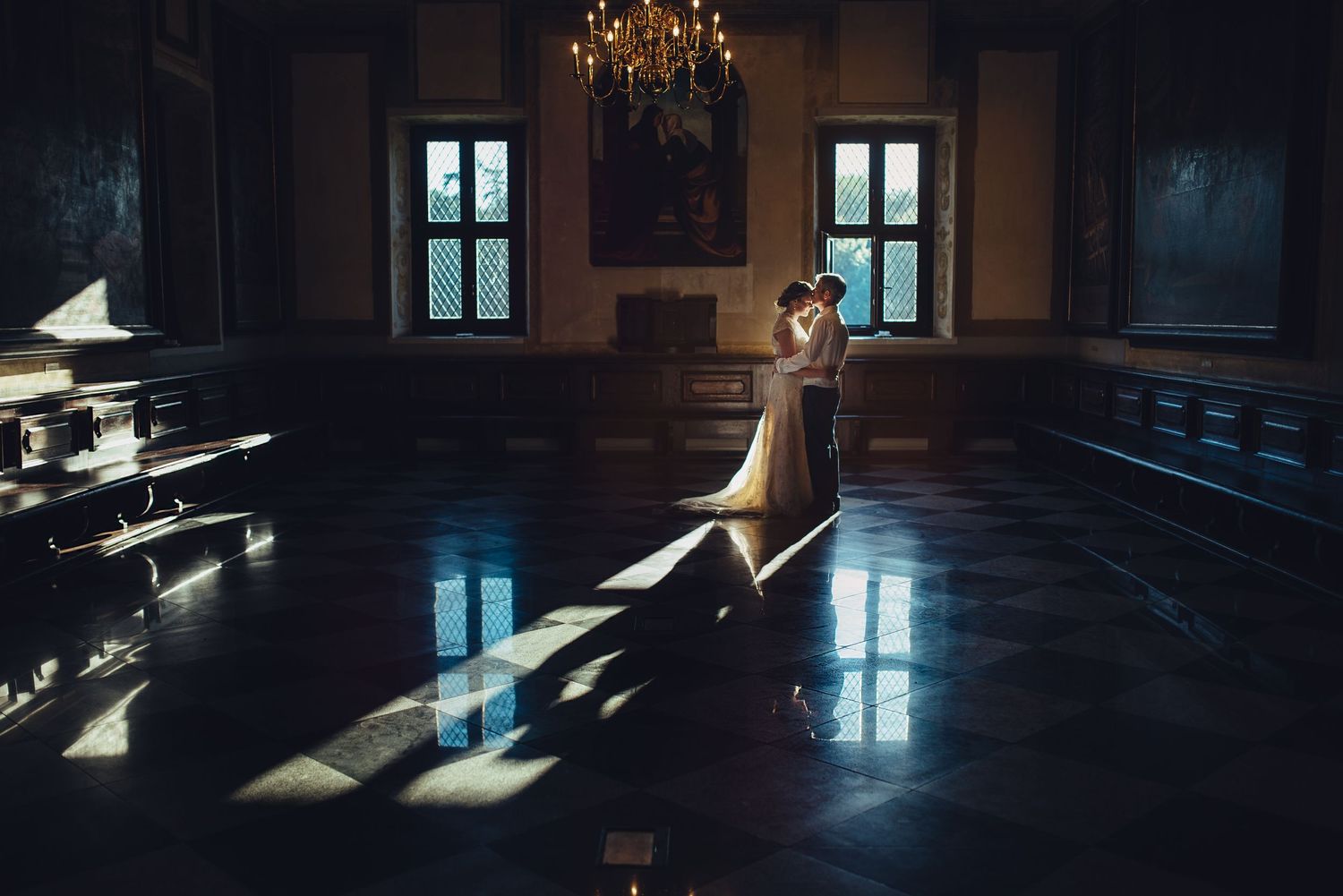 Dramatic shadows cast through windows onto dark hardwood floors in an elegant room with a chandelier.