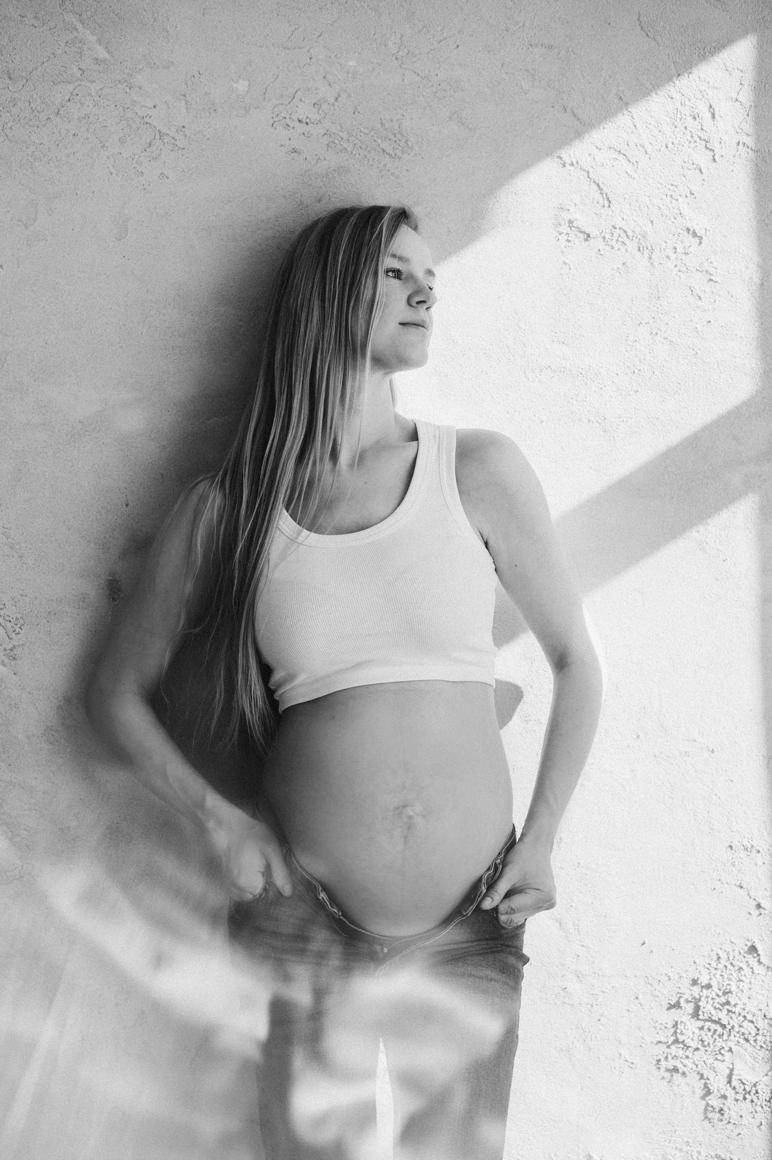 Minimalist maternity portrait in white against textured wall with dramatic lighting.