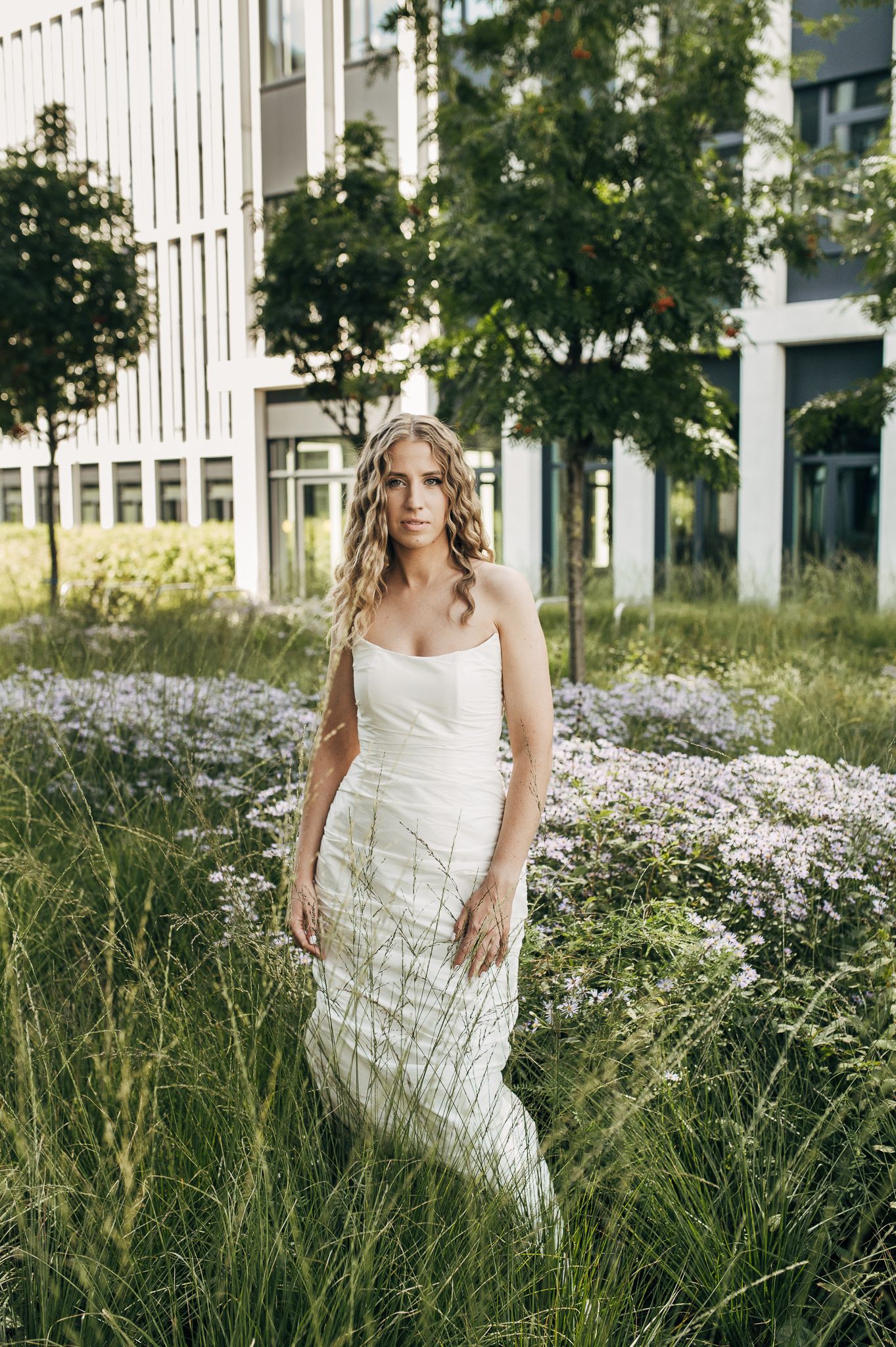 Elegant white wedding dress photographed in a garden setting with blooming white flowers and modern architecture.