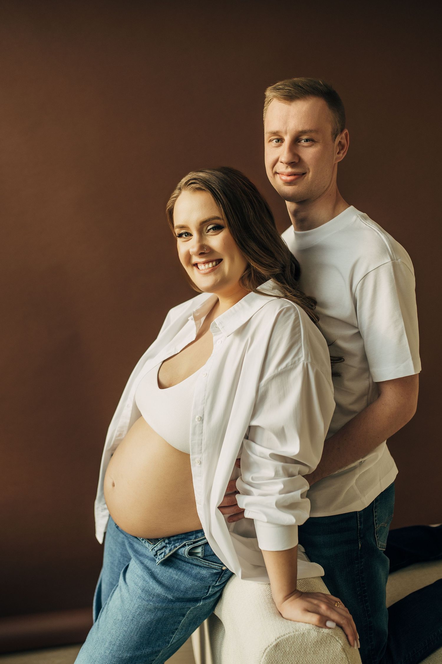 Expecting couple poses together in casual attire during maternity photoshoot with warm brown backdrop.