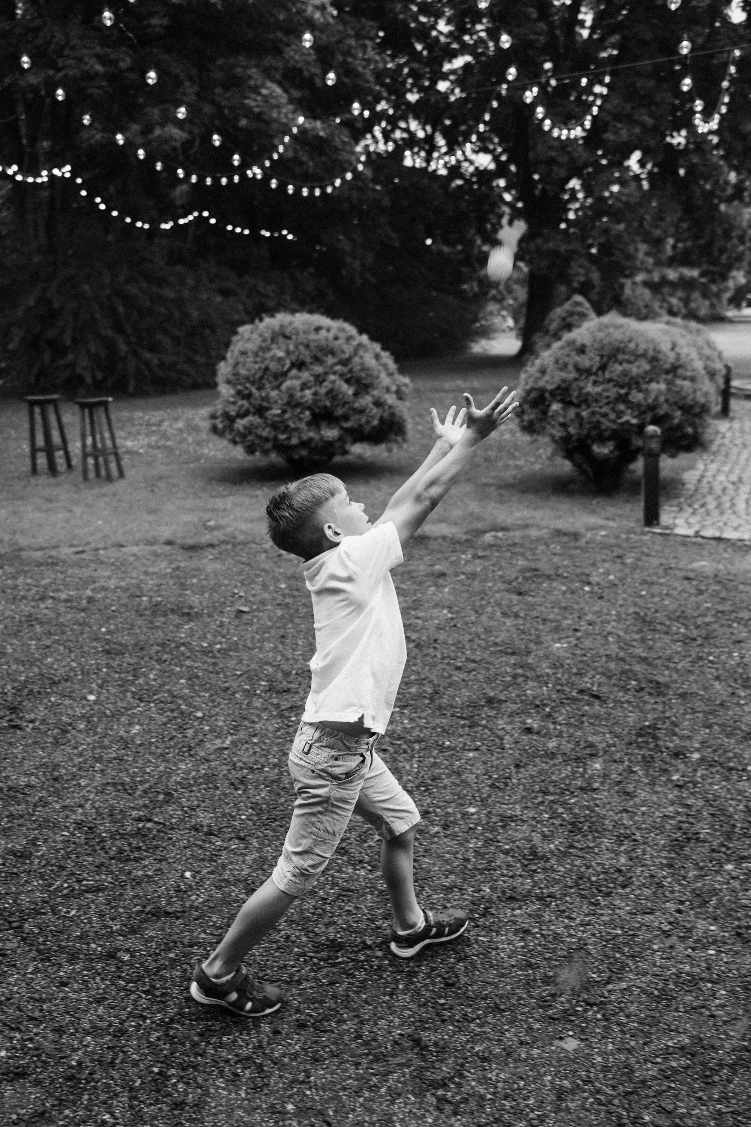 Black and white photo of a boy reaching up toward string lights in a garden park.