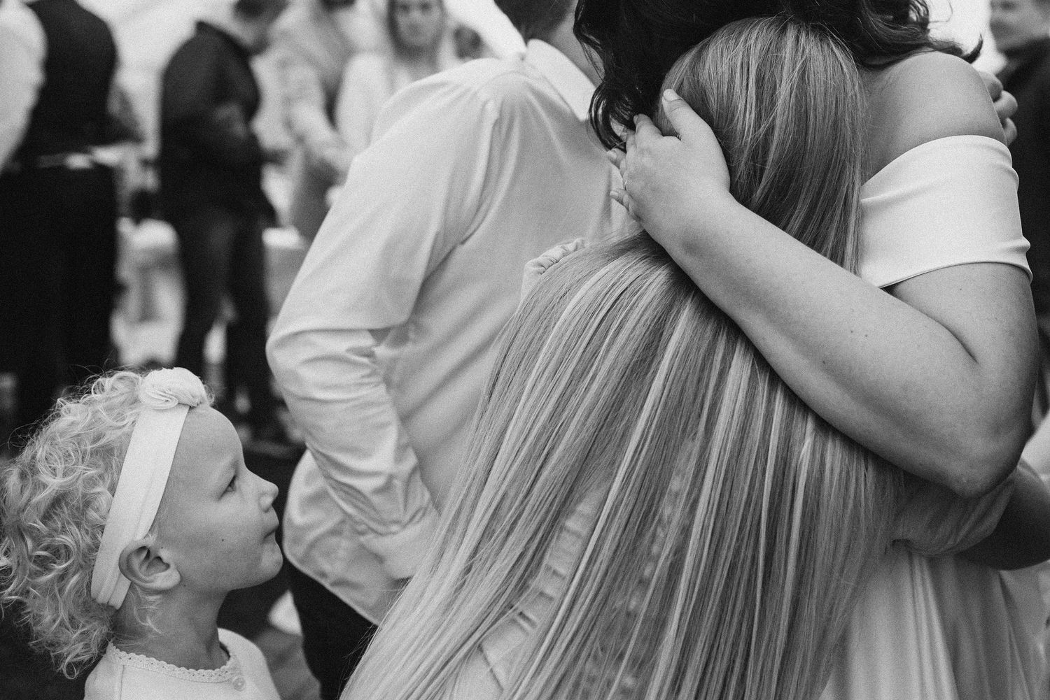 Black and white photo of a curly-haired flower girl hugging a bride at a wedding reception.