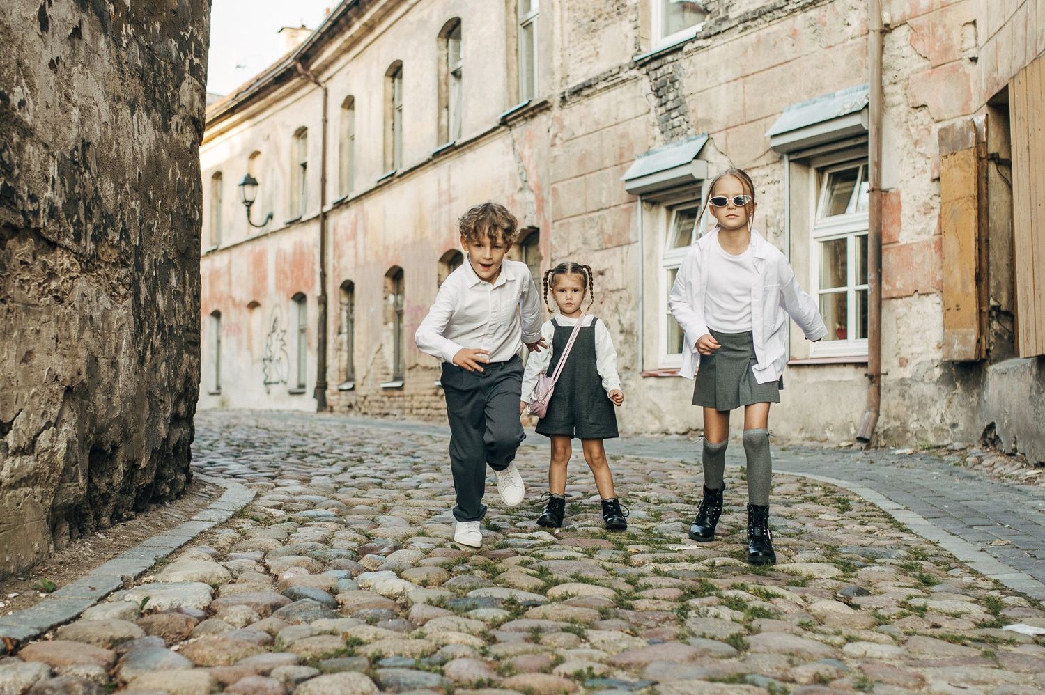 Three children in vintage-style clothing walk together on a cobblestone street in an old European town.