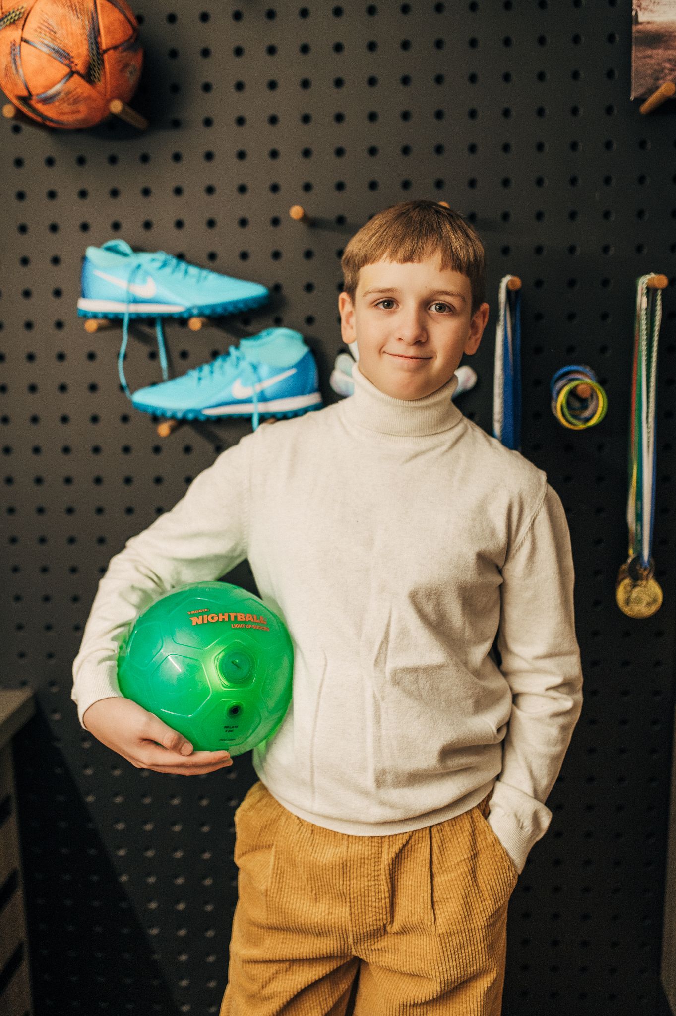Child in cream sweater and corduroy pants posing with sports equipment against dark pegboard wall.