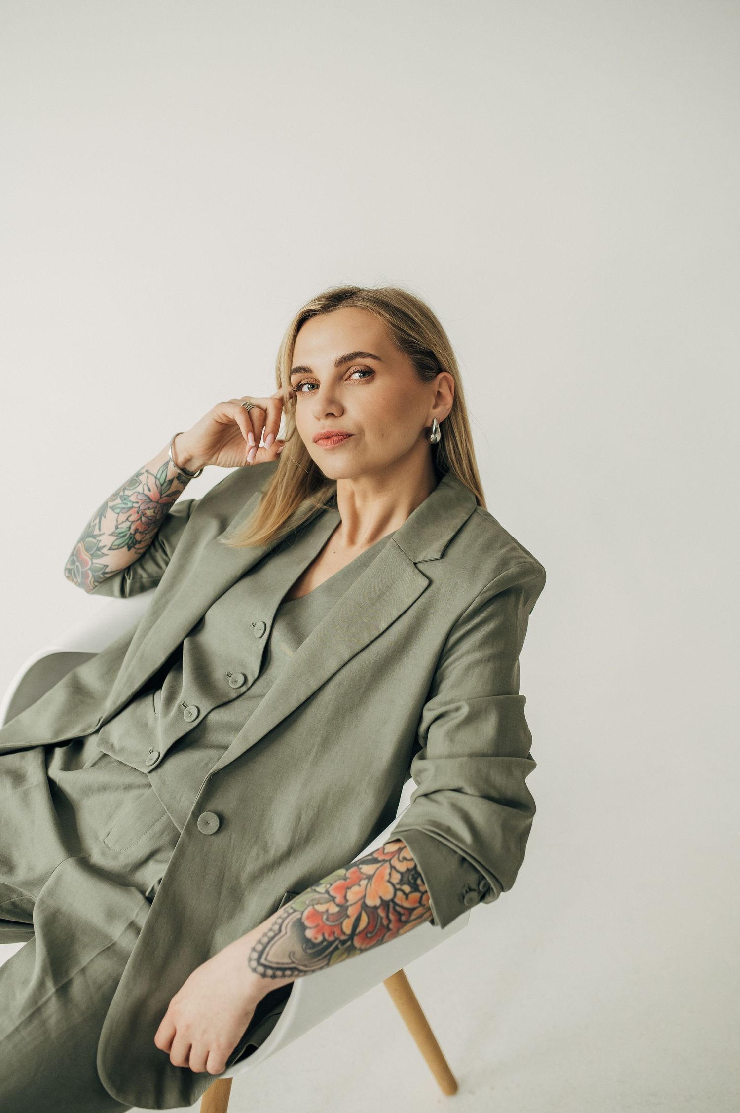 Woman with tattoos poses in sage green blazer against white wall.