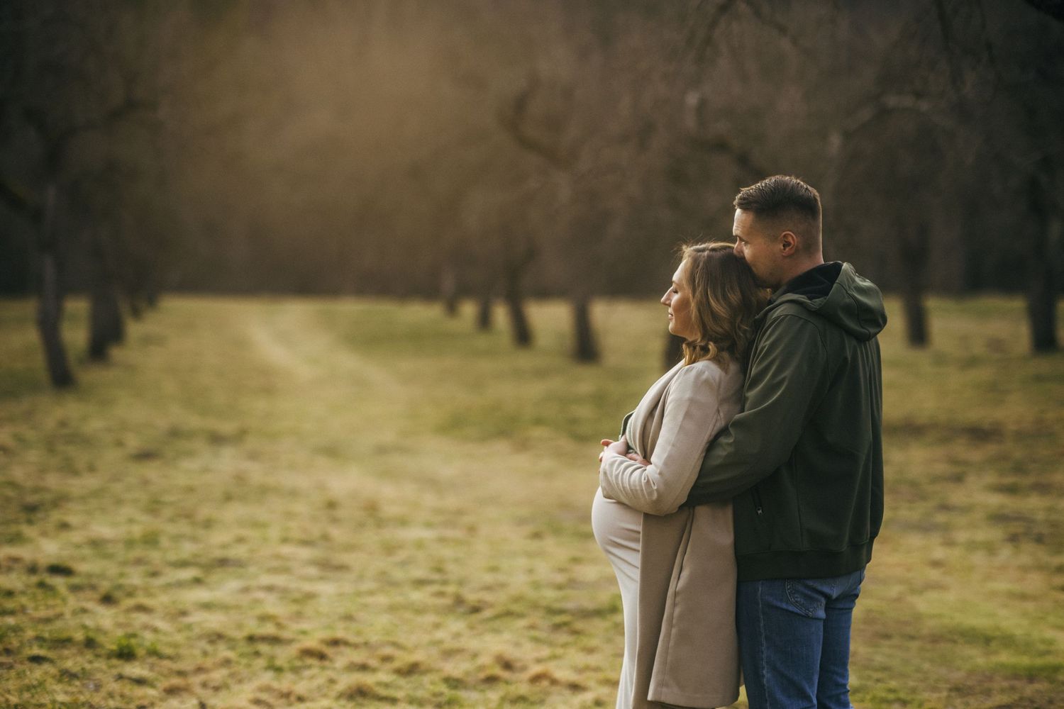 Couple embracing outdoors in autumn orchard, pregnant woman and man sharing tender moment on scenic tree-lined path.