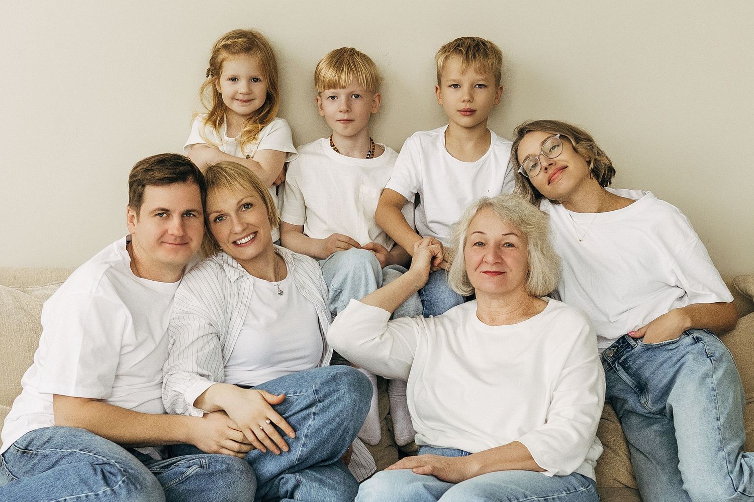 Multi-generational family wearing white tops and blue jeans poses together on beige couch for portrait.