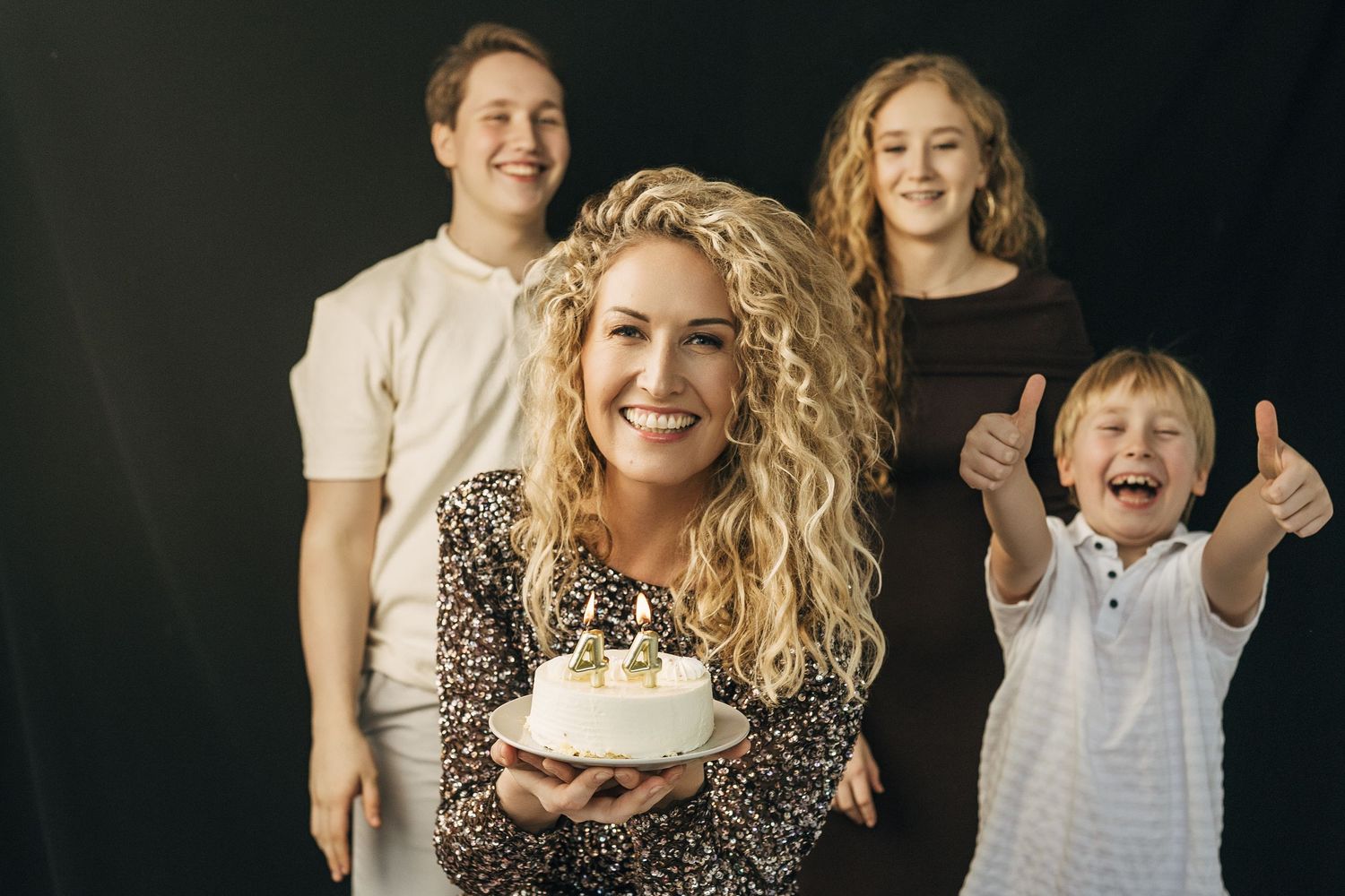 Woman holding birthday cake smiles with family of three behind her against dark background.