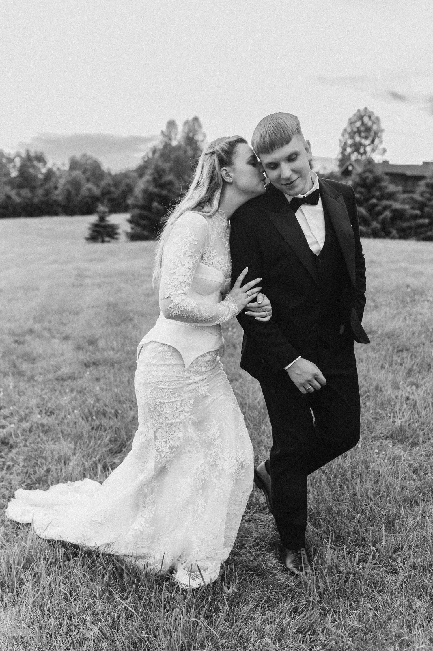 Black and white wedding photo of a couple embracing on a grassy hillside with trees in the background.