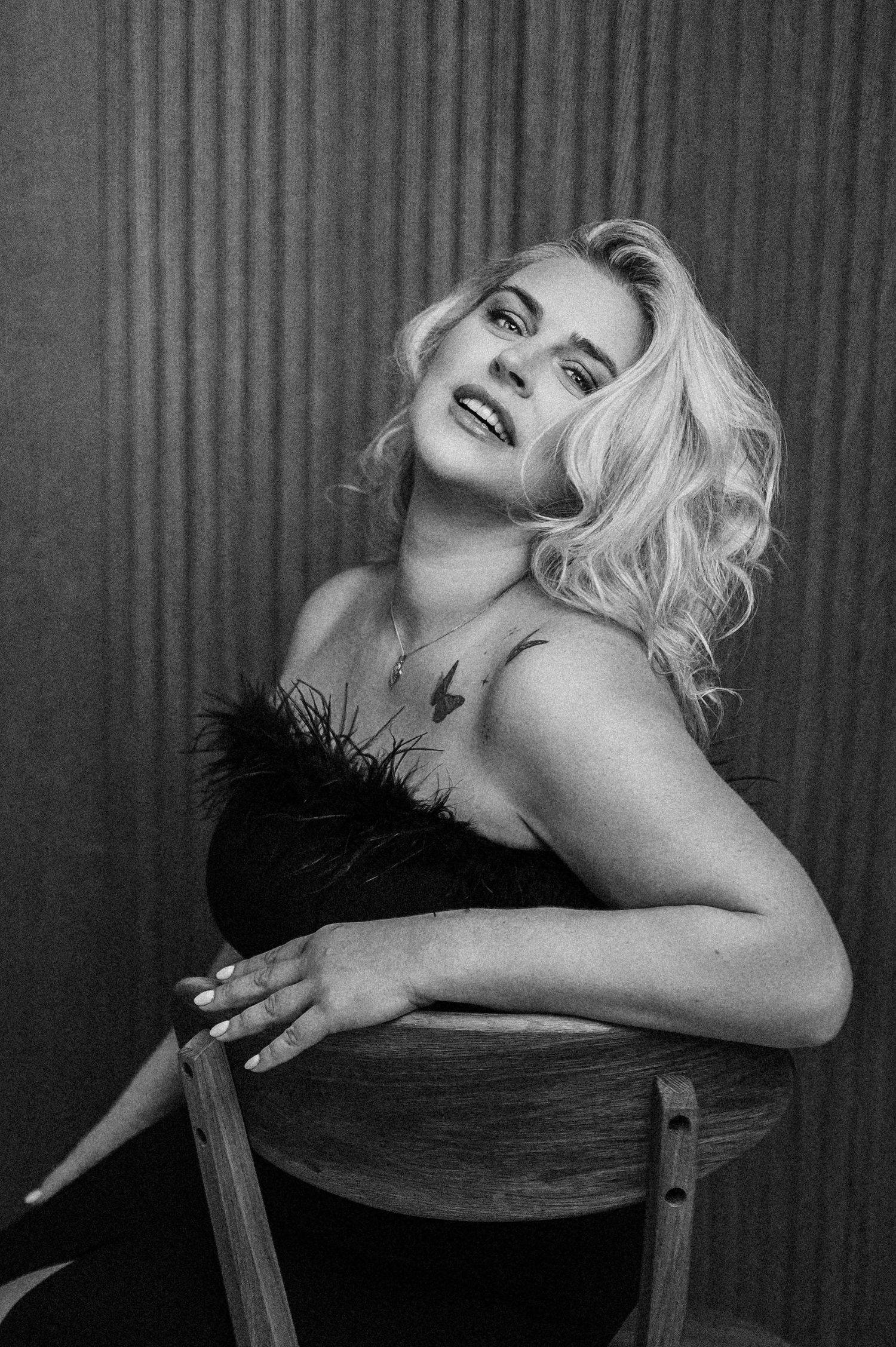 Black and white portrait of a blonde woman in a feathered top, seated on a stool, looking upward confidently.