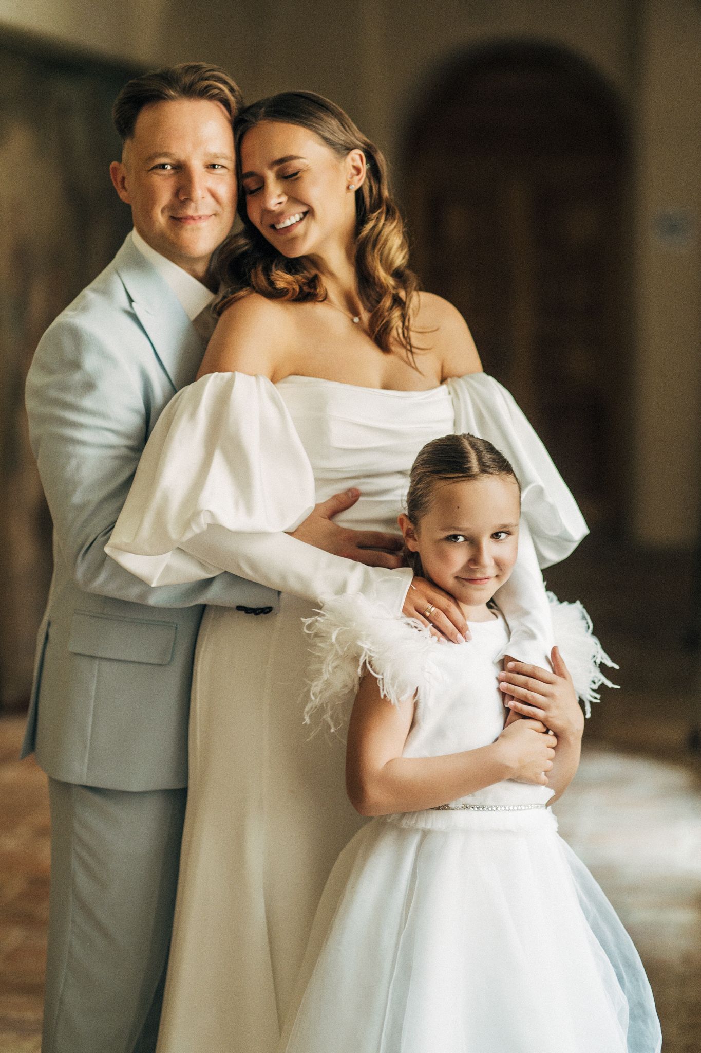Happy wedding family portrait with groom in gray suit, bride in off-shoulder white gown, and young daughter.