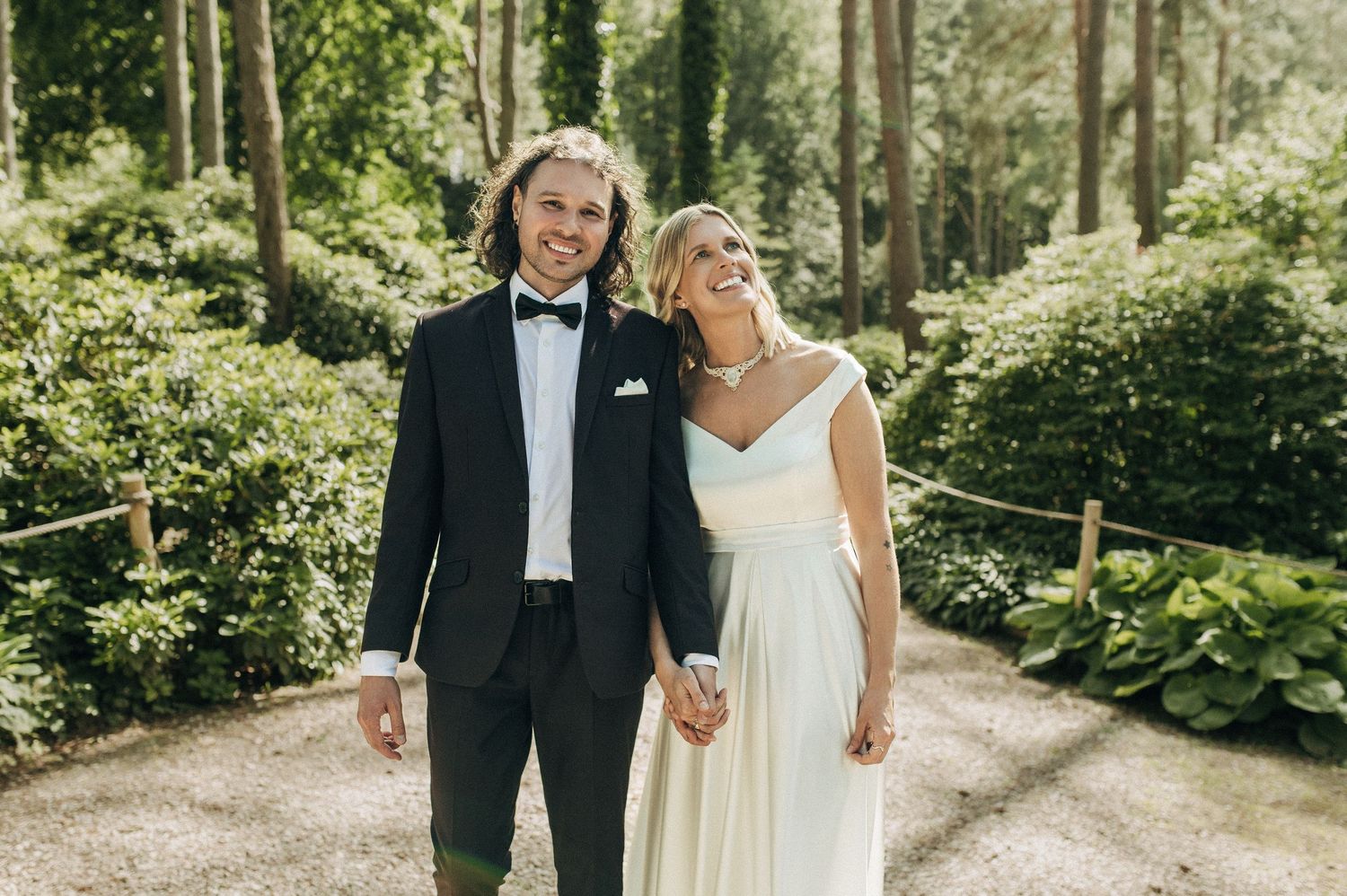 A couple in wedding attire walks together through a sunlit garden path surrounded by lush greenery.