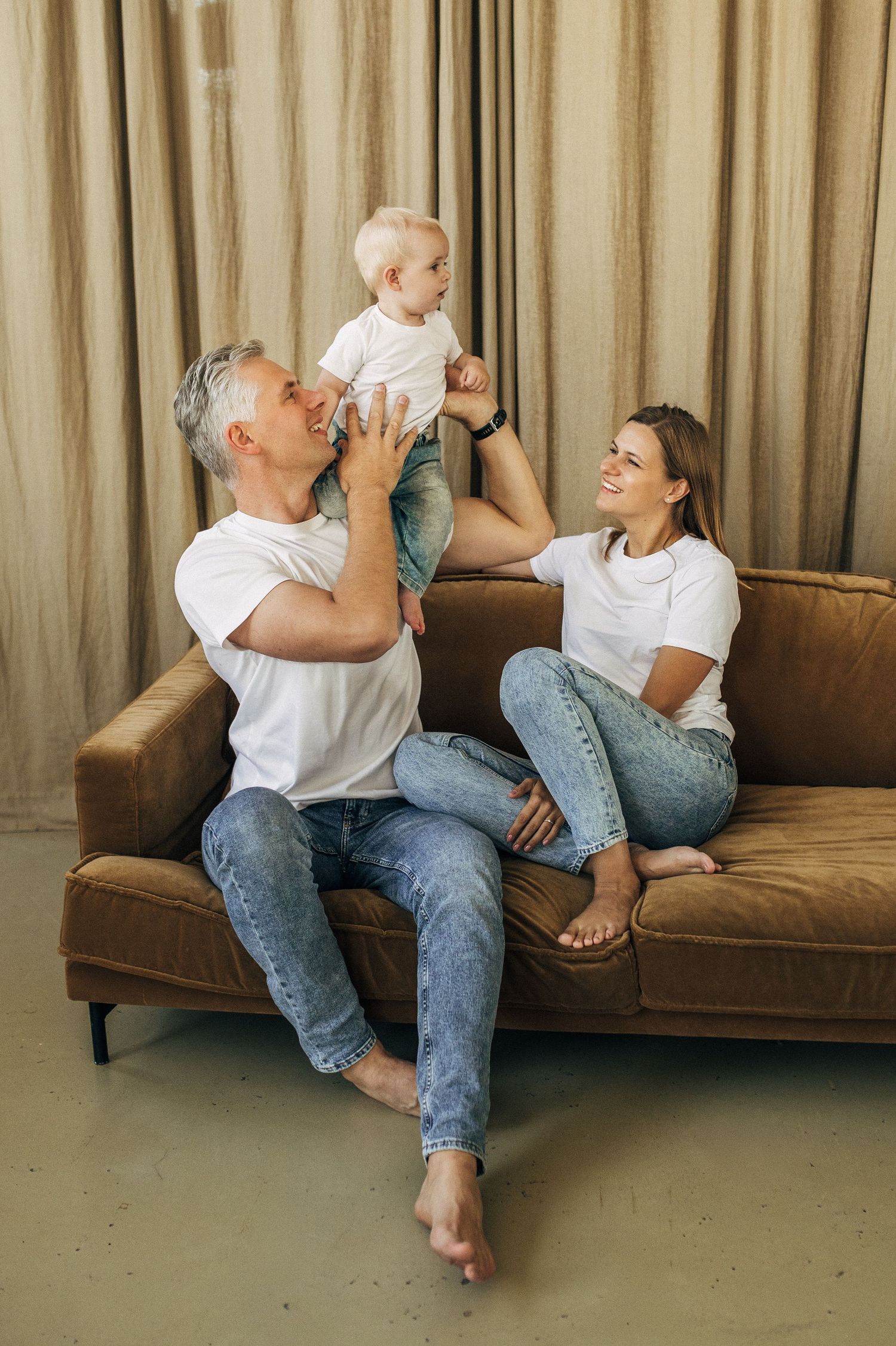 Family spending quality time together on brown couch, playing and laughing in casual white t-shirts and jeans.