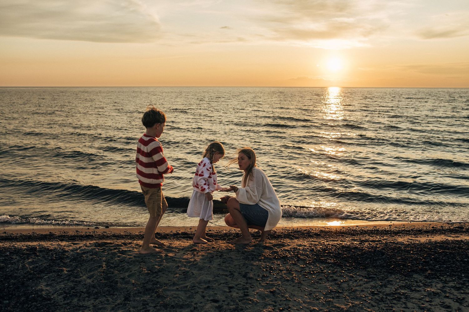 A mother crouches on a sandy beach with two children at golden sunset, waves gently lapping the shore.
