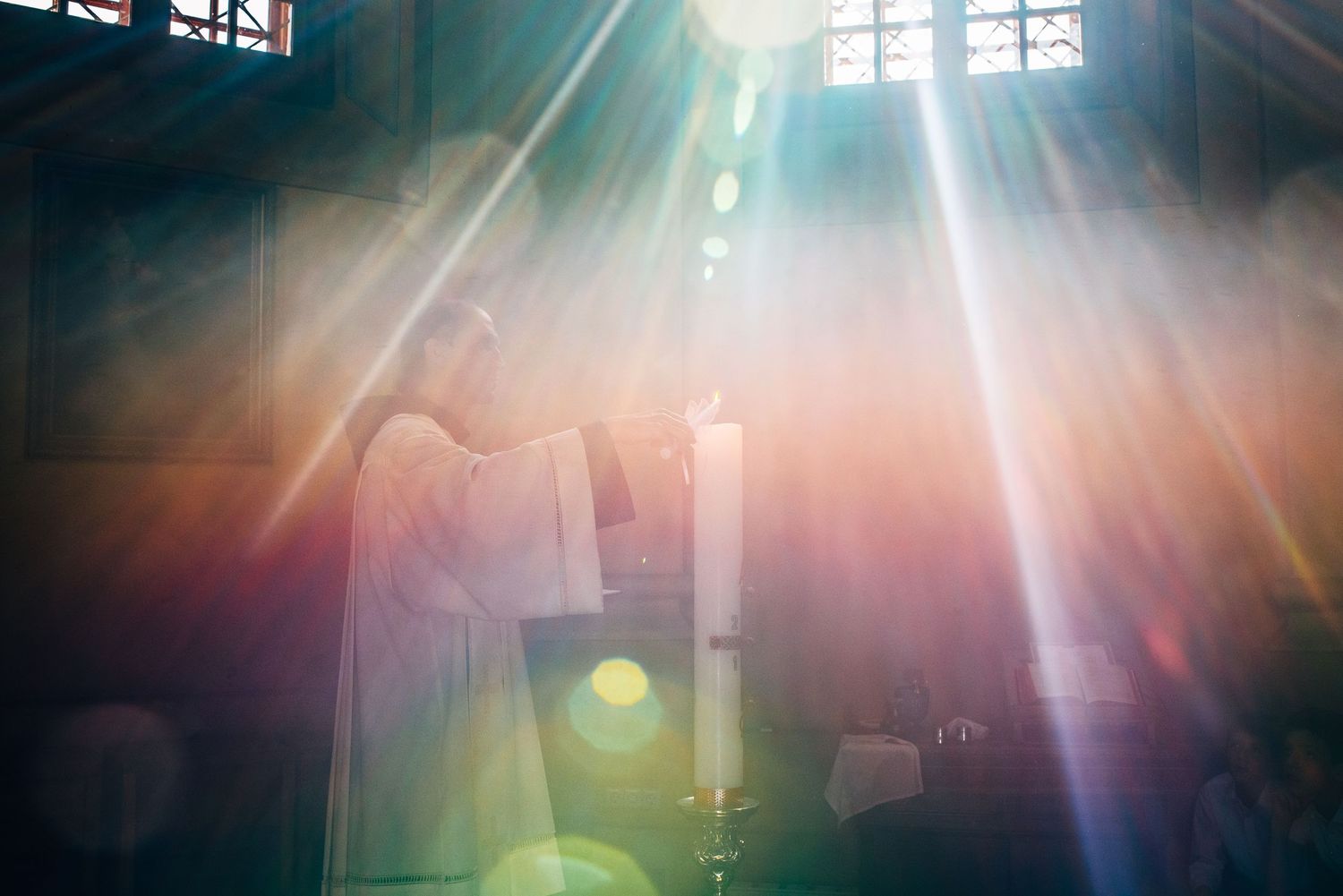 Ethereal church scene with dramatic sunbeams illuminating a figure holding a candle at altar.
