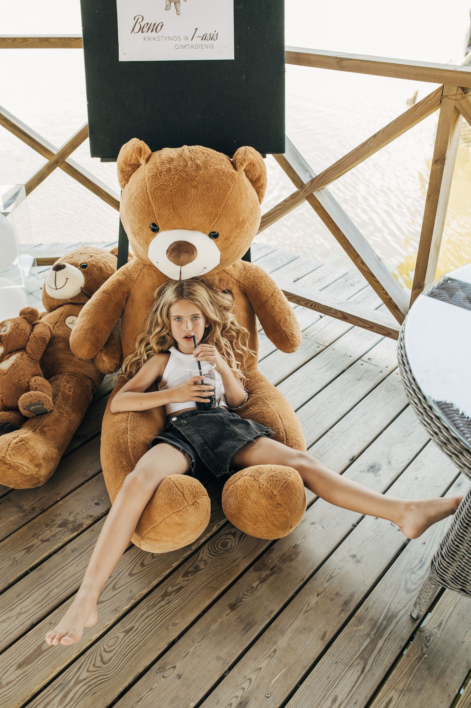 Young girl sitting against a giant brown teddy bear on a wooden deck, holding a drink, surrounded by more stuffed animals.