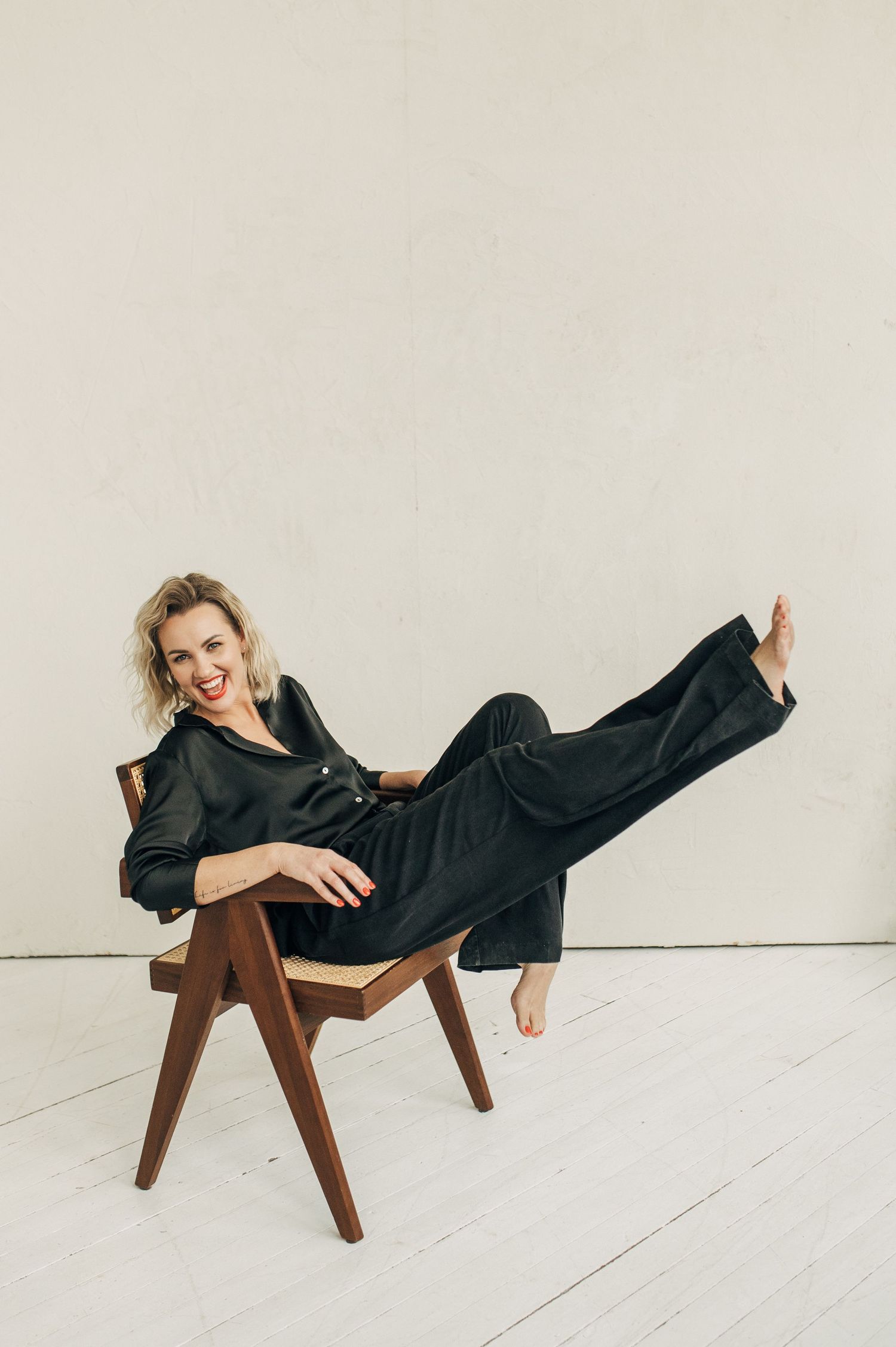 Fashion photoshoot series of model reclining on wooden chair in black outfit against white background.