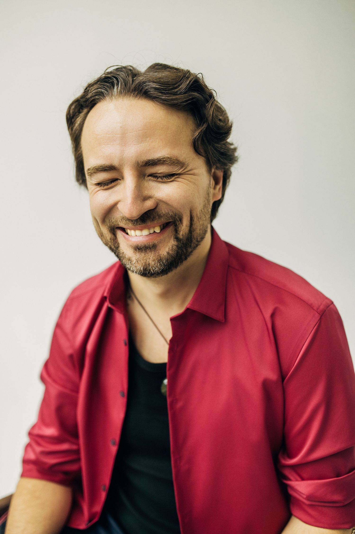 Smiling man with beard wearing red shirt laughing joyfully against white background.
