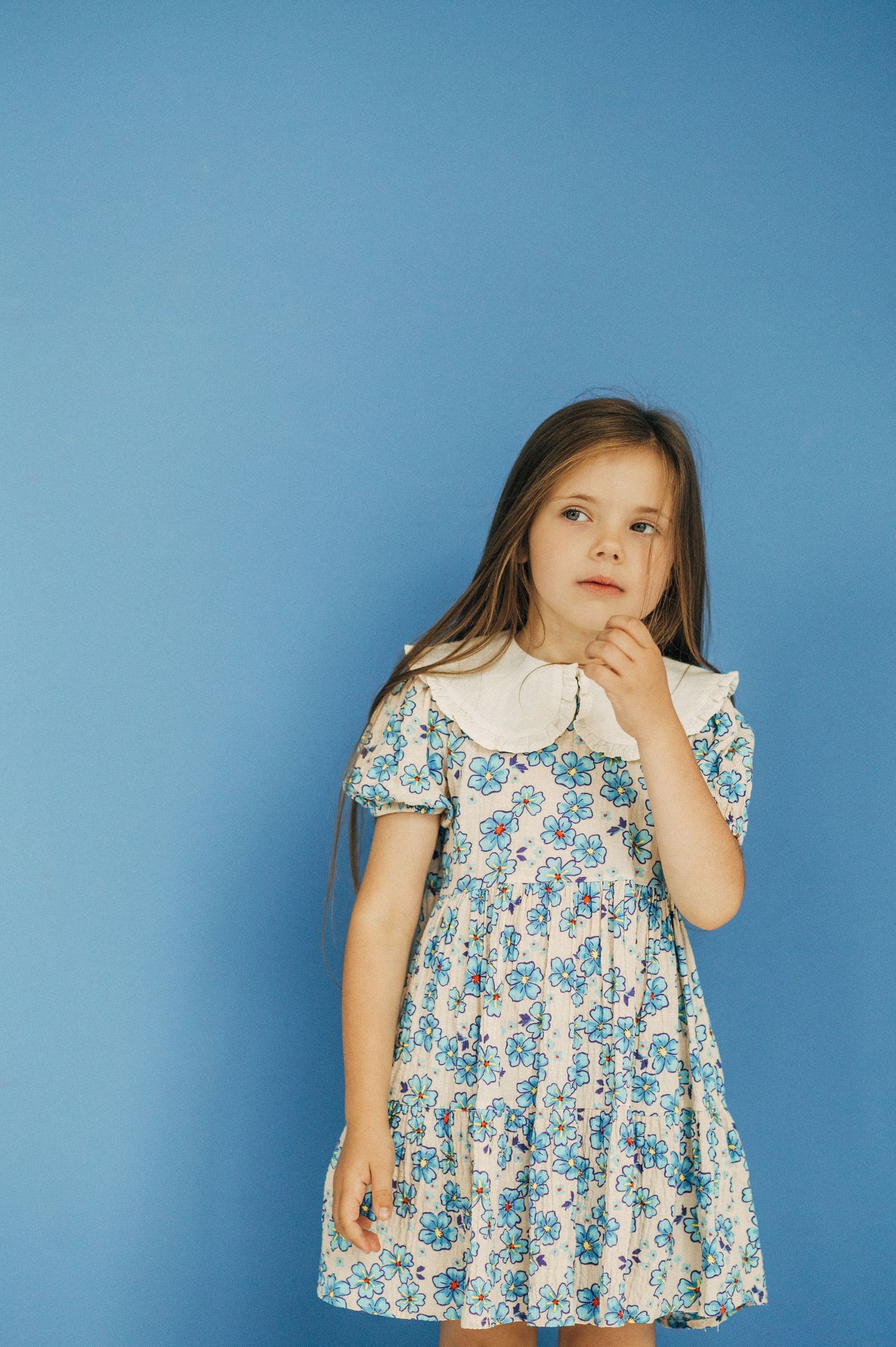 Child in floral dress posing expressively against blue studio backdrop.
