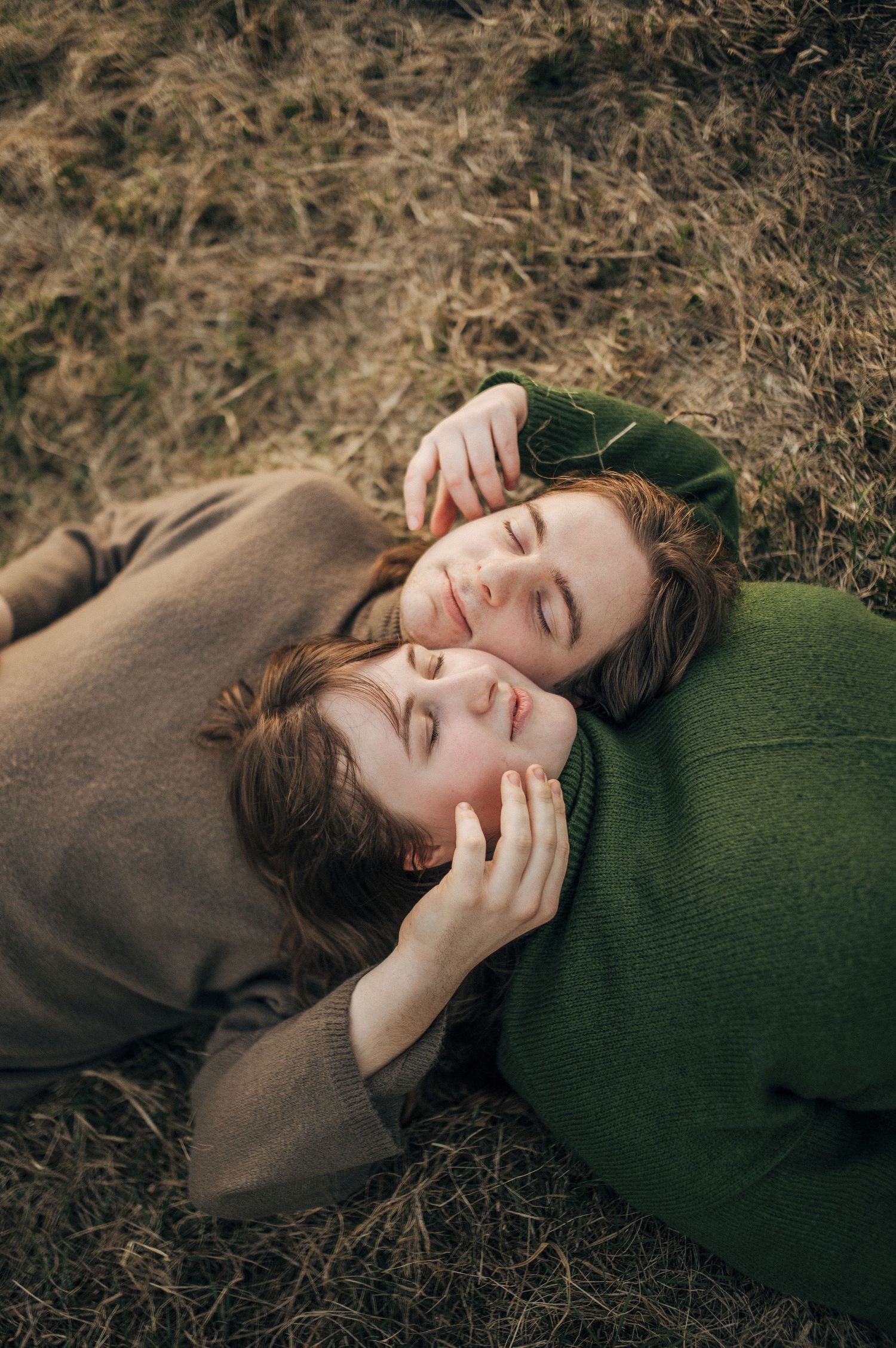 Two people lying in dried grass wearing cozy sweaters on an autumn day.