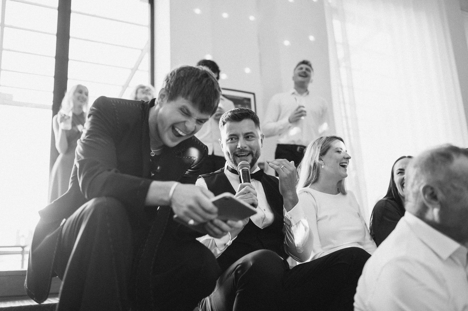Black and white photo of guests laughing and enjoying a lively social gathering or wedding reception indoors.