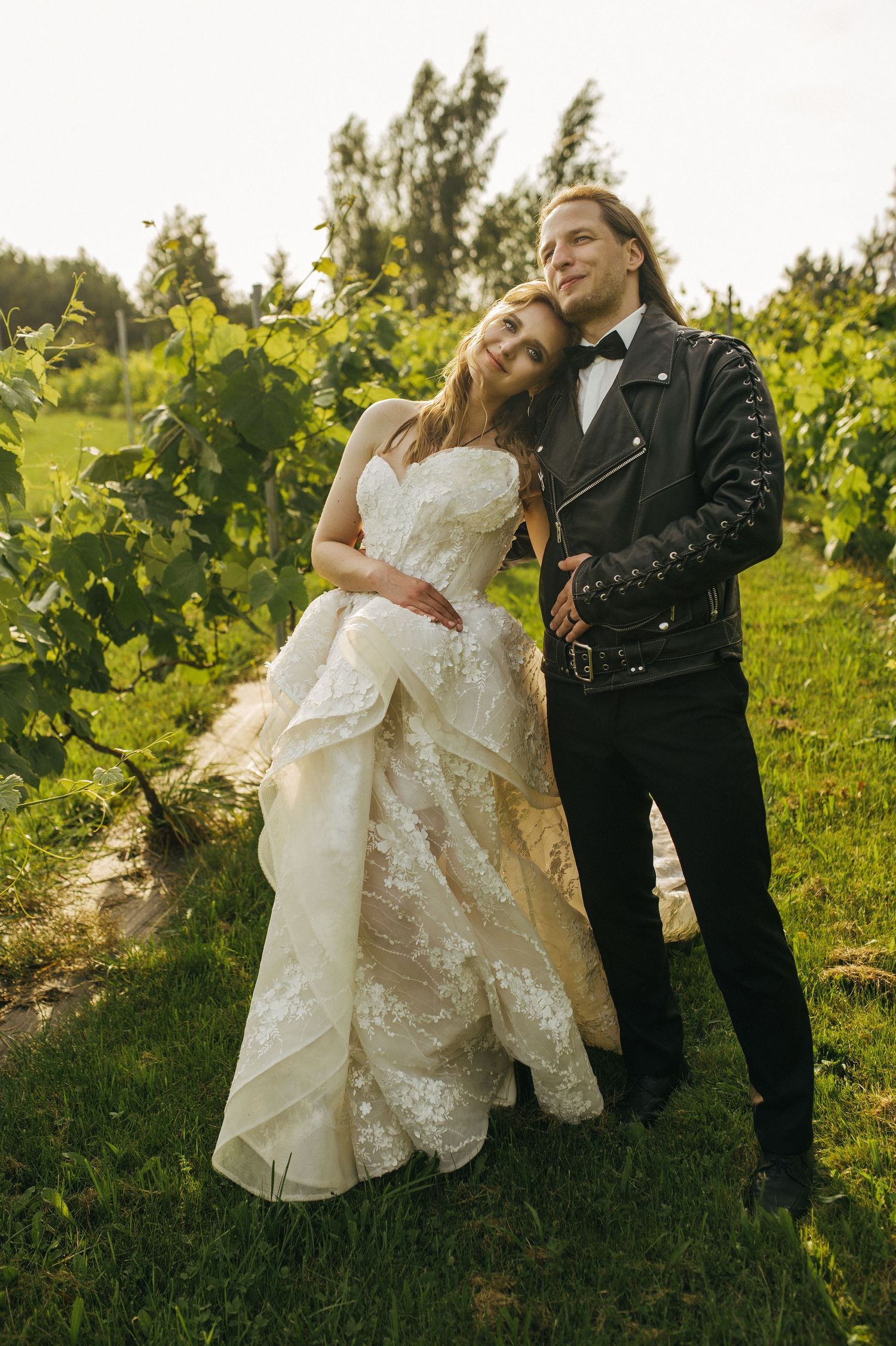 Bride in lace gown and groom in leather jacket embrace in a lush vineyard during their wedding photo session.