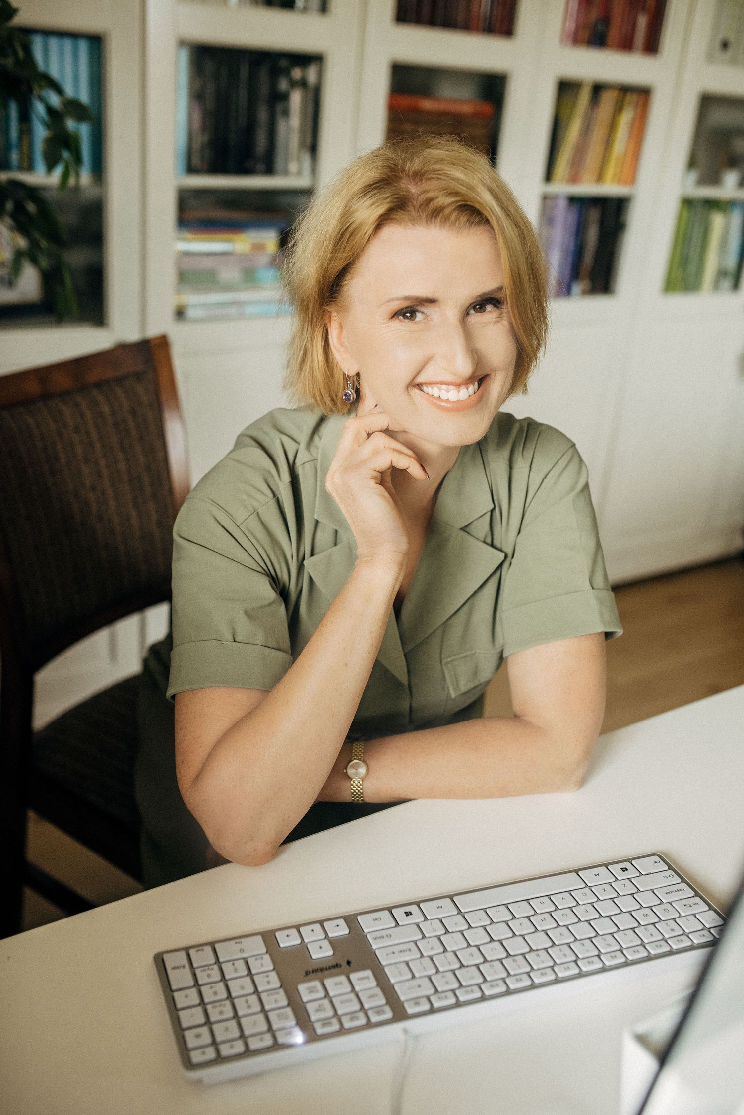 Professional wearing olive green shirt smiles while working at desk with Apple keyboard in home office setting.
