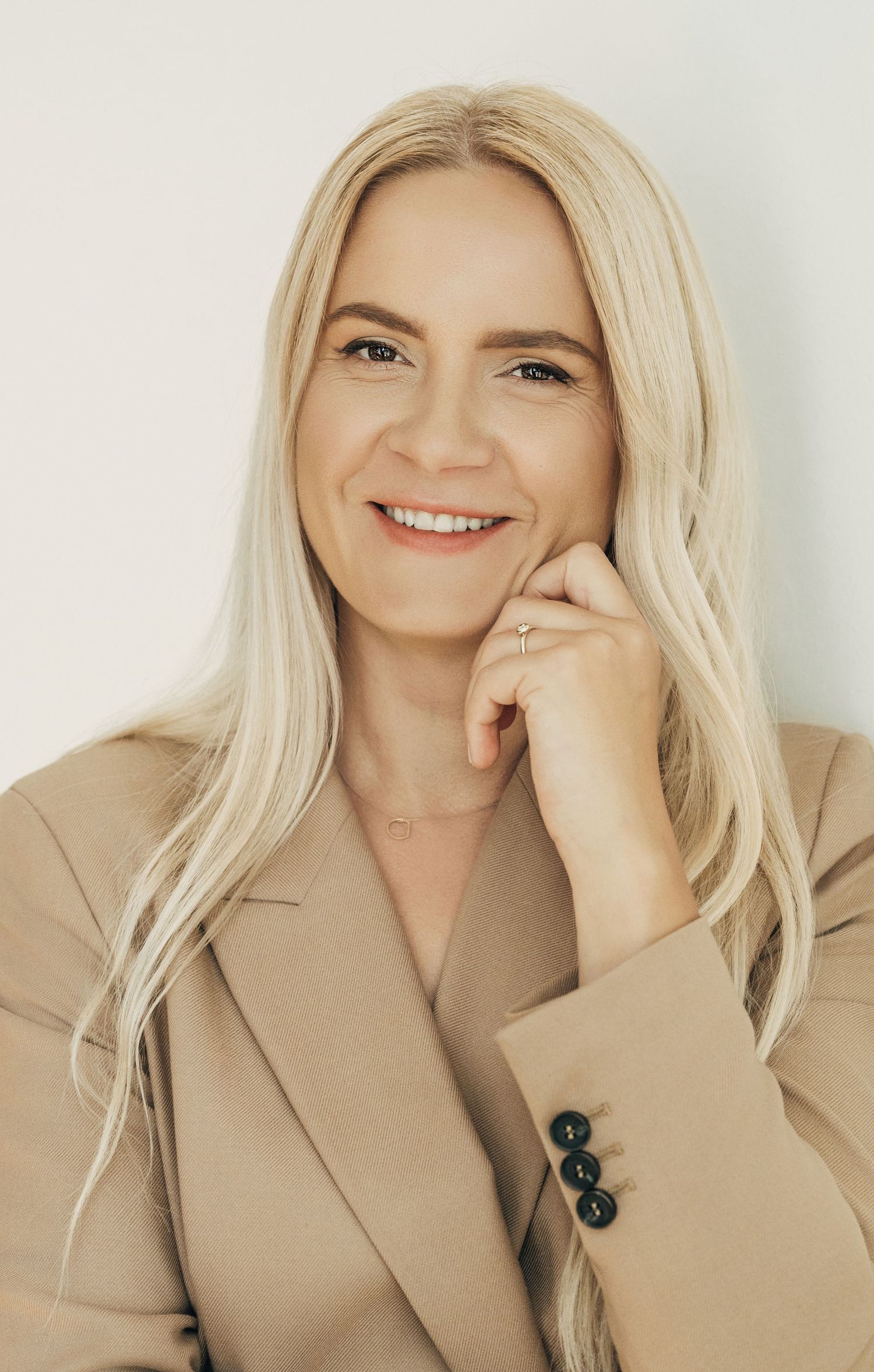 Professional headshot of someone in beige blazer against white background with warm expression.