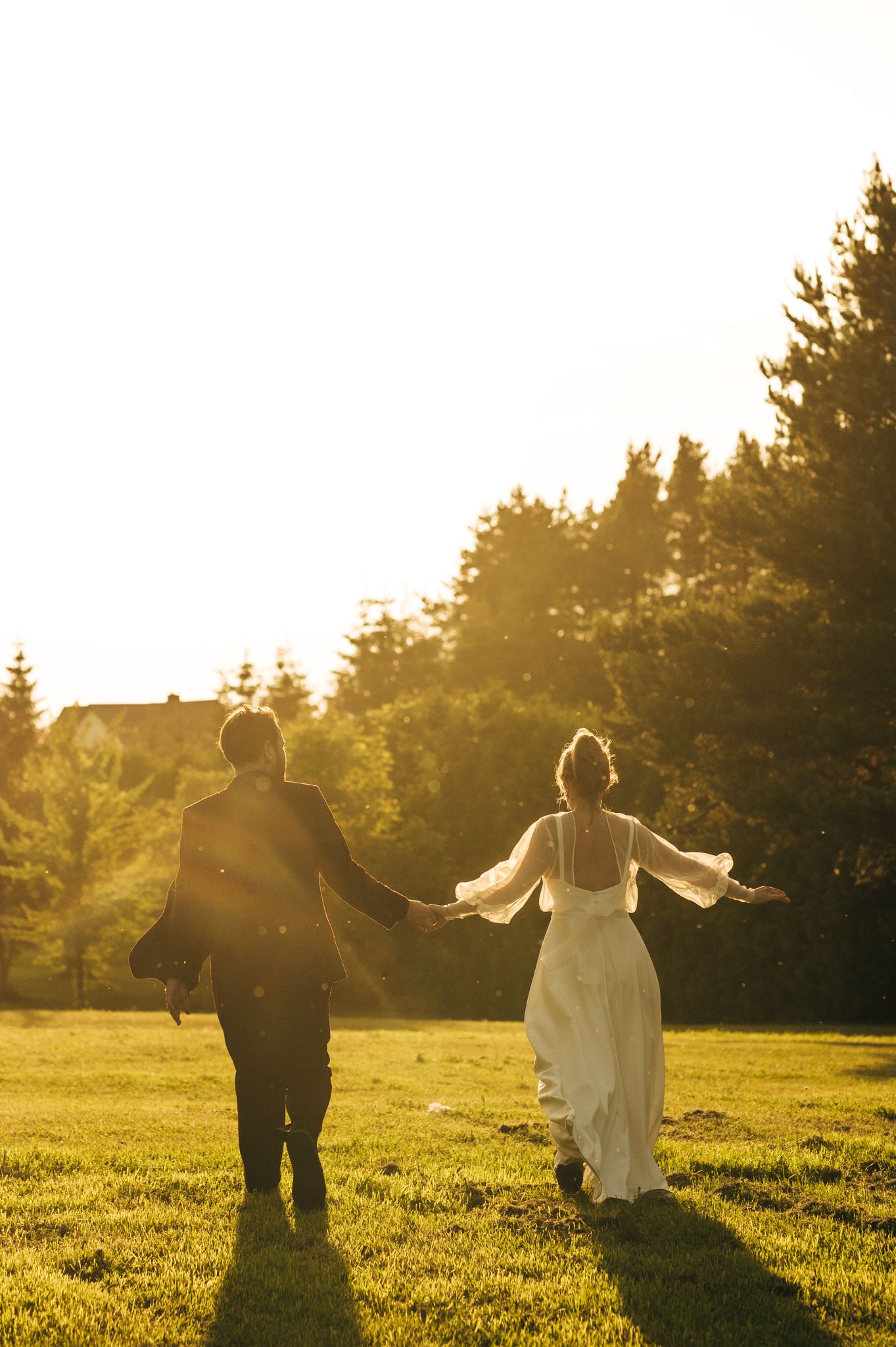 A couple runs through a sunlit meadow at golden hour with their arms outstretched.