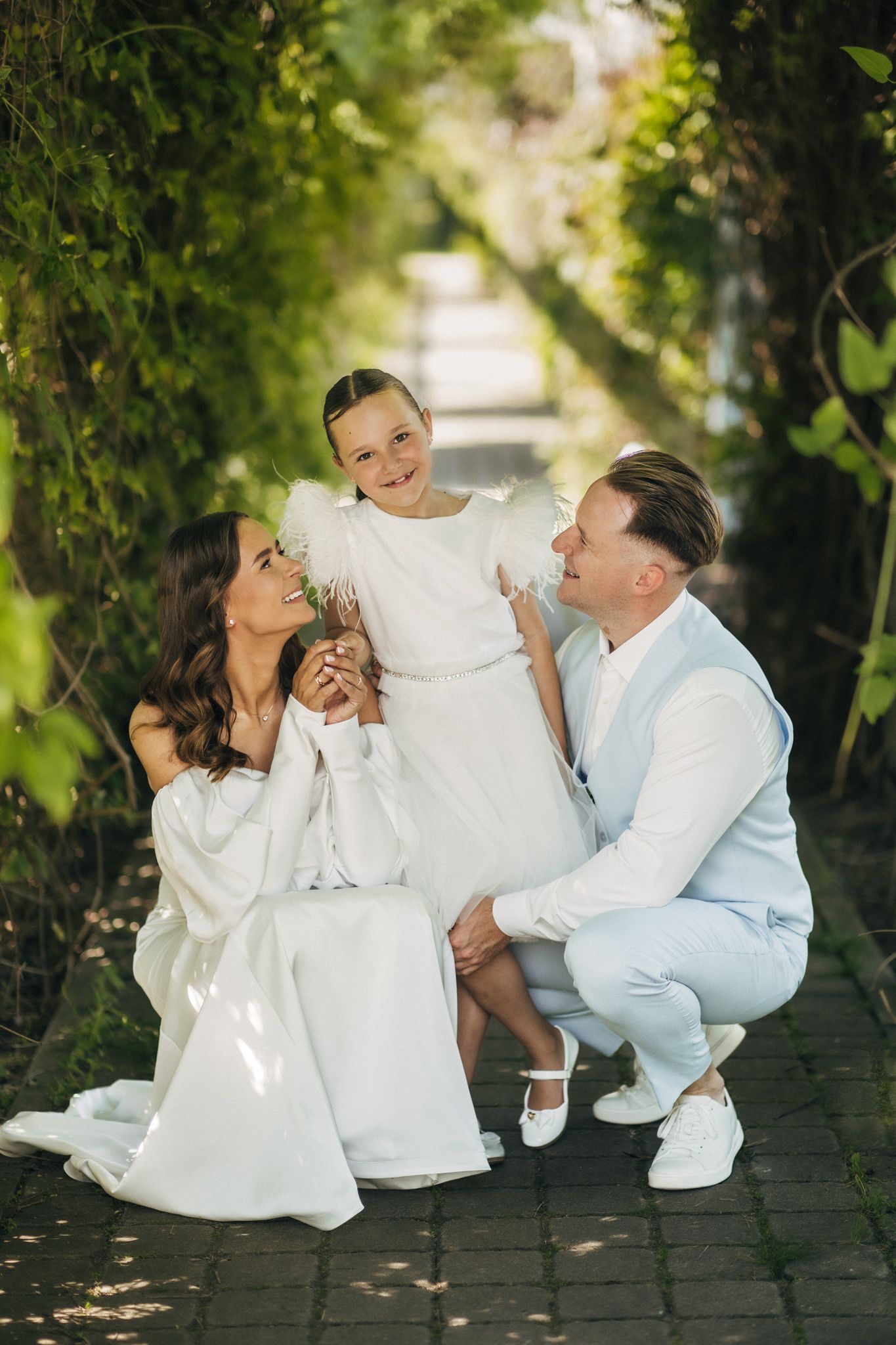 Happy family of three in white outfits posing in lush green garden pathway.